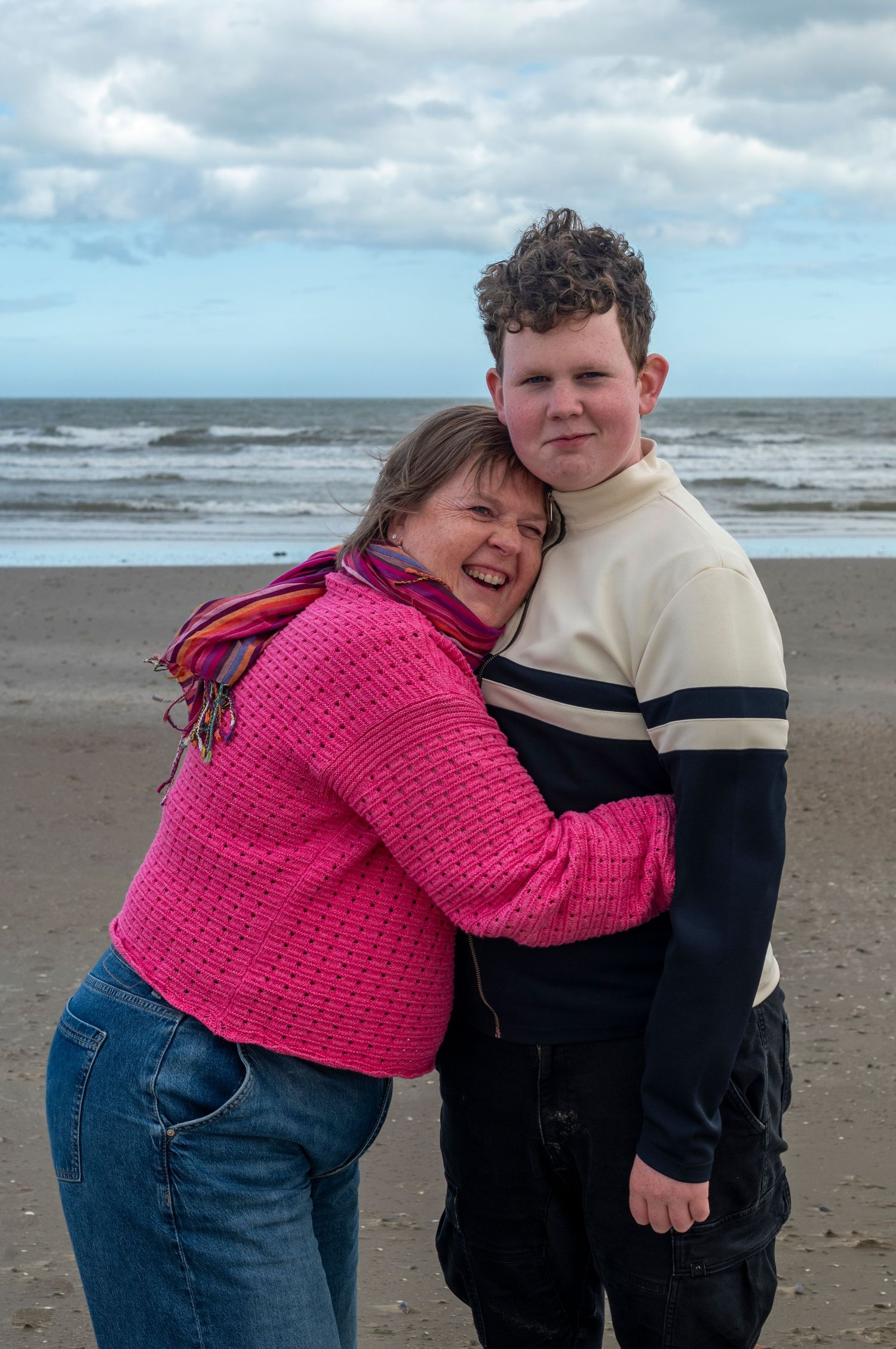 A woman in a pink sweater hugs a teenage boy on a sandy beach with ocean waves and cloudy sky behind them.