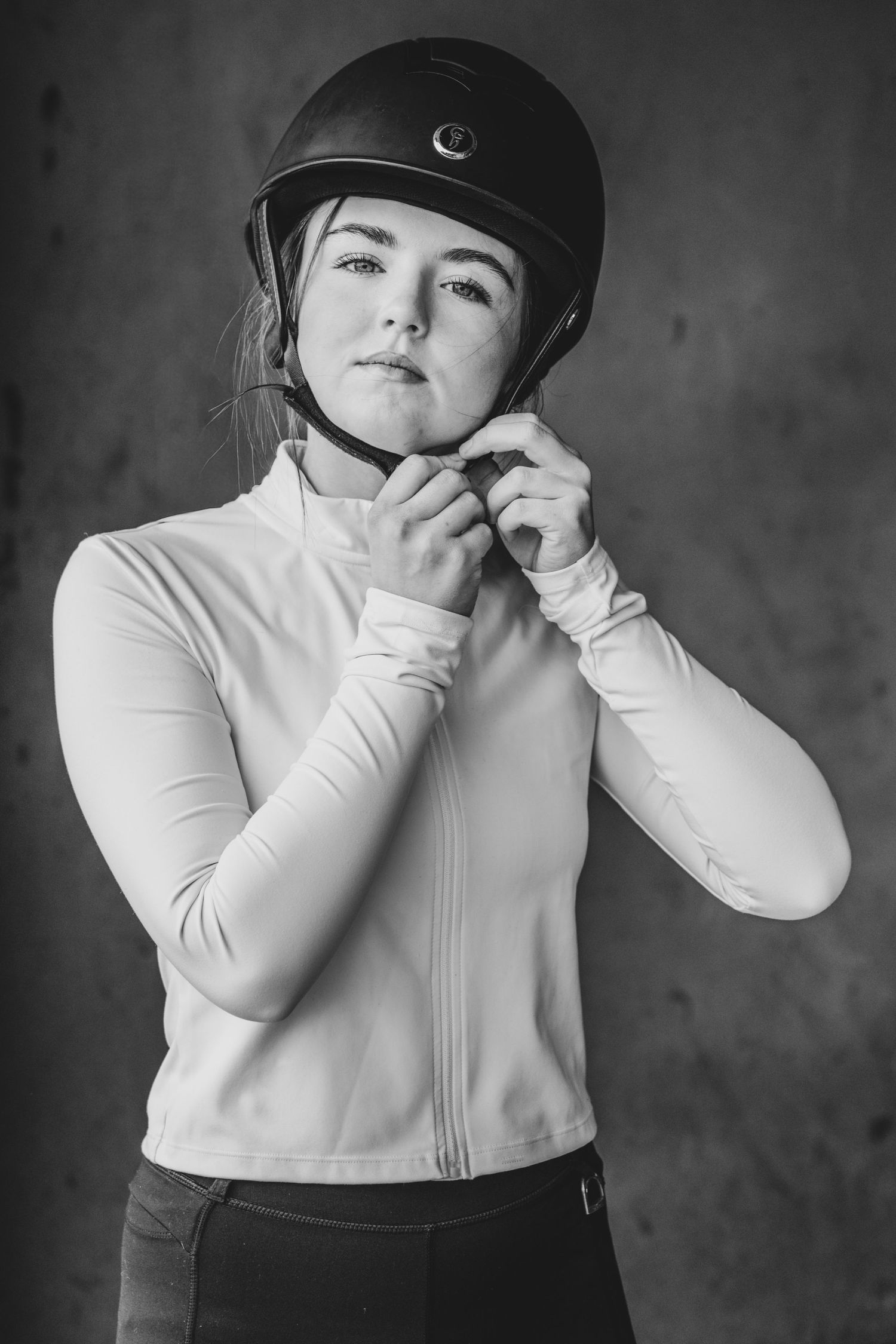 Black and white photo of a young woman fastening a black equestrian helmet while wearing a white long-sleeve jacket.