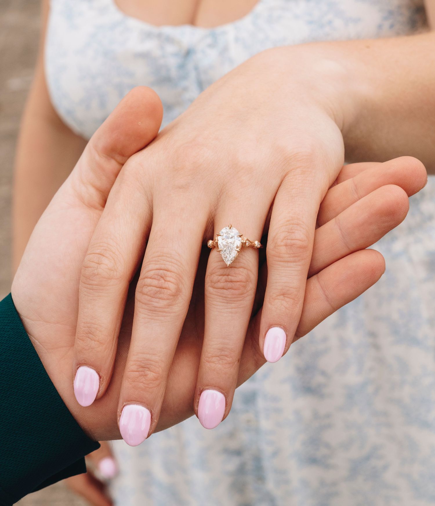 Close-up of an engagement ring with a pear-shaped diamond on a gold band on a womans finger with pink nails.