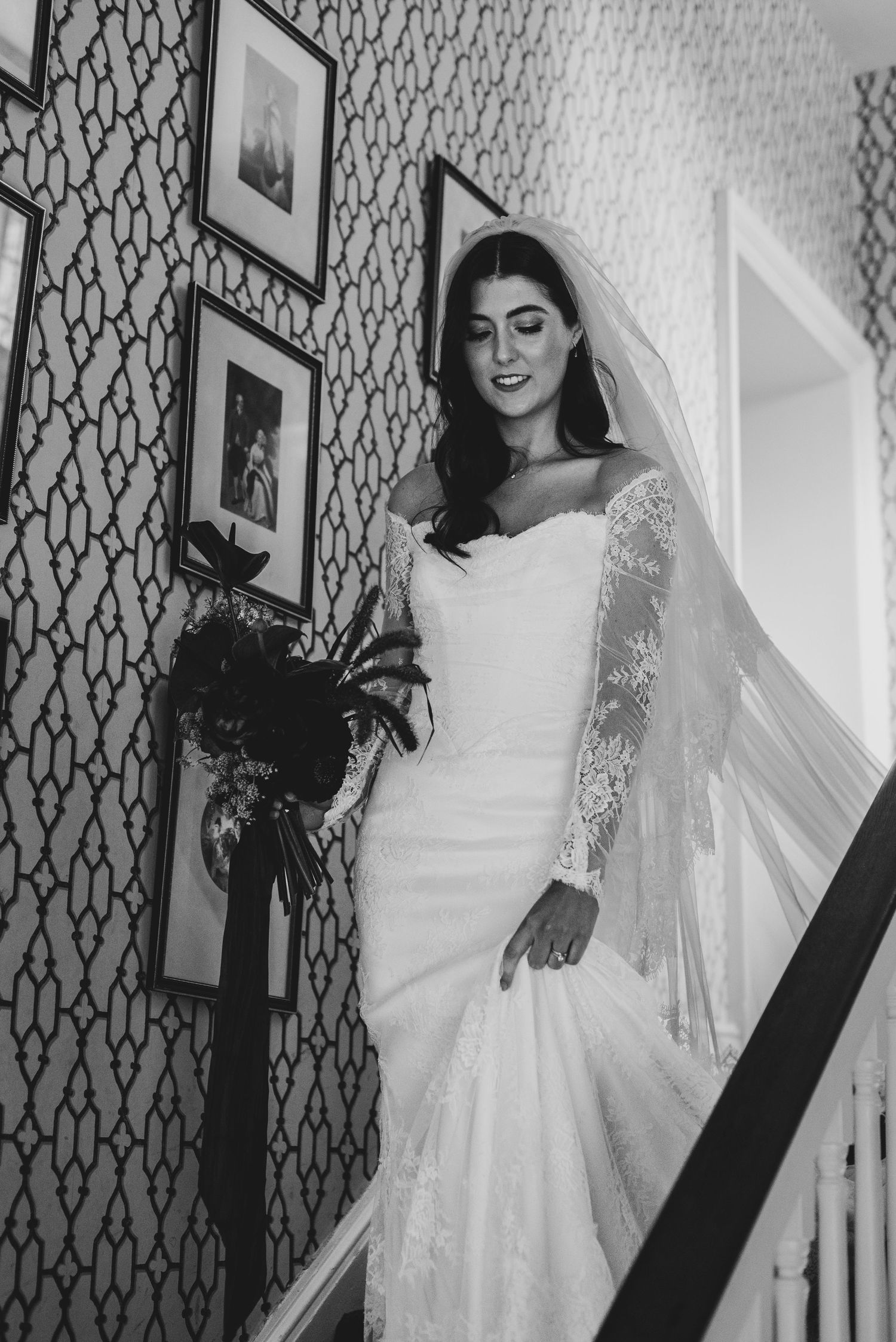 Bride in lace mermaid gown and veil descends staircase holding dark floral bouquet in black and white photo.