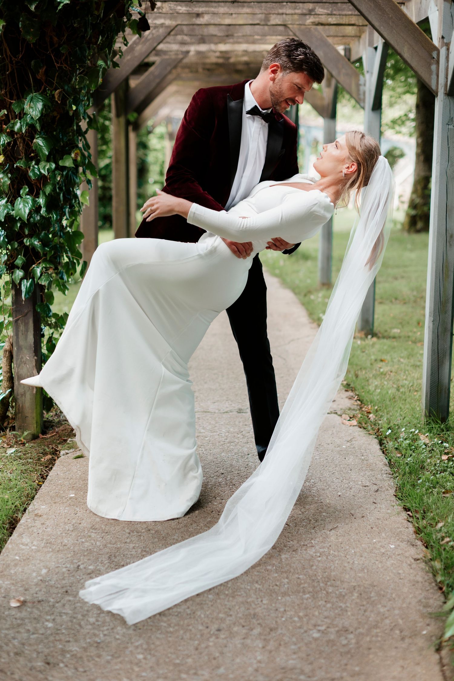 Groom in burgundy velvet tuxedo dips bride in white gown under ivy-covered pergola.