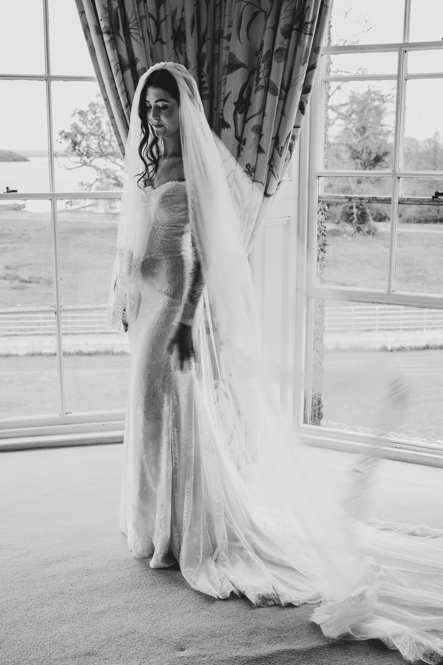 Black and white photo of a bride in a lace gown and flowing veil standing by large windows.