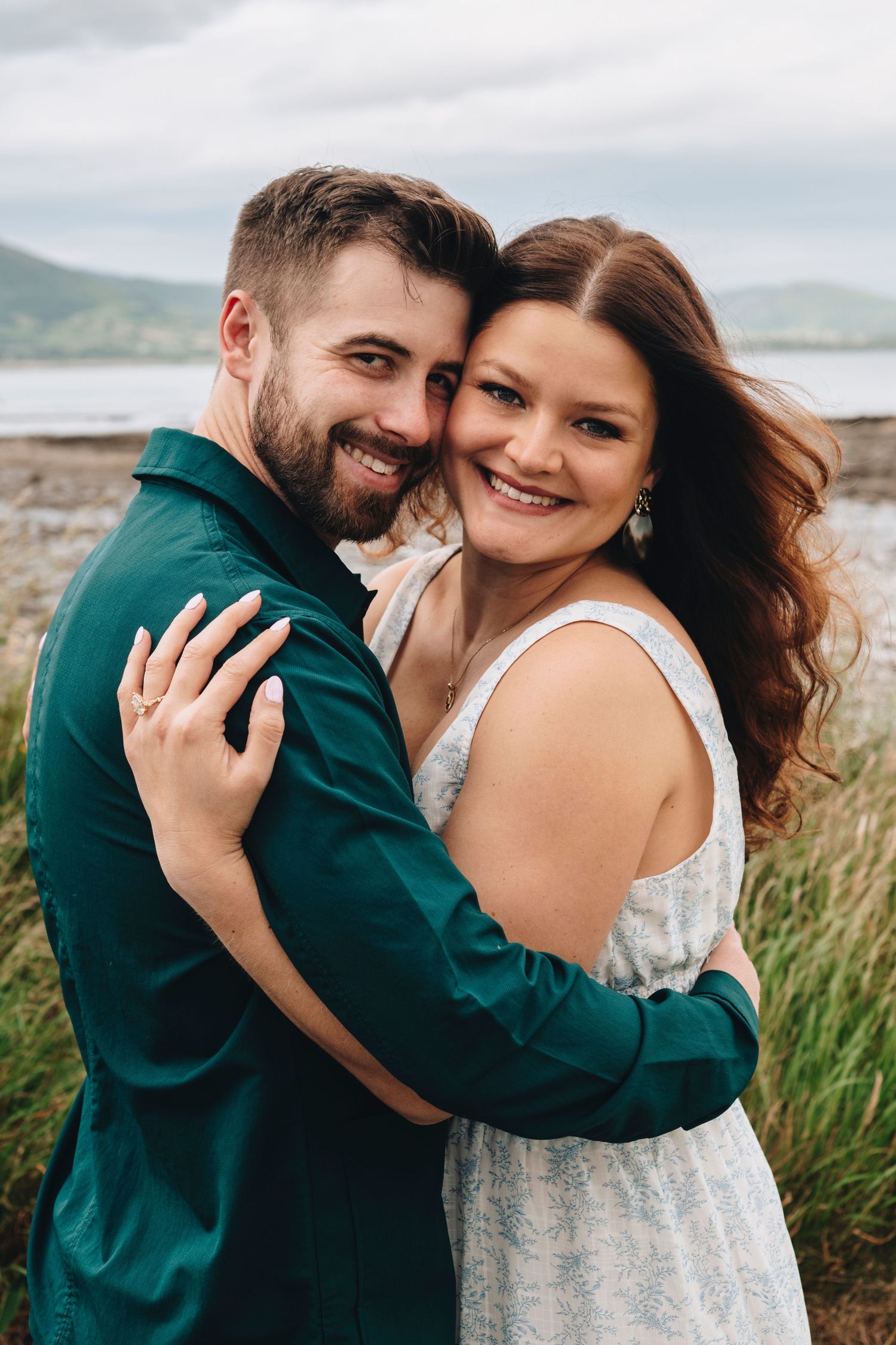 Smiling couple embracing outdoors near a lake with mountains, woman in white dress, man in green shirt.