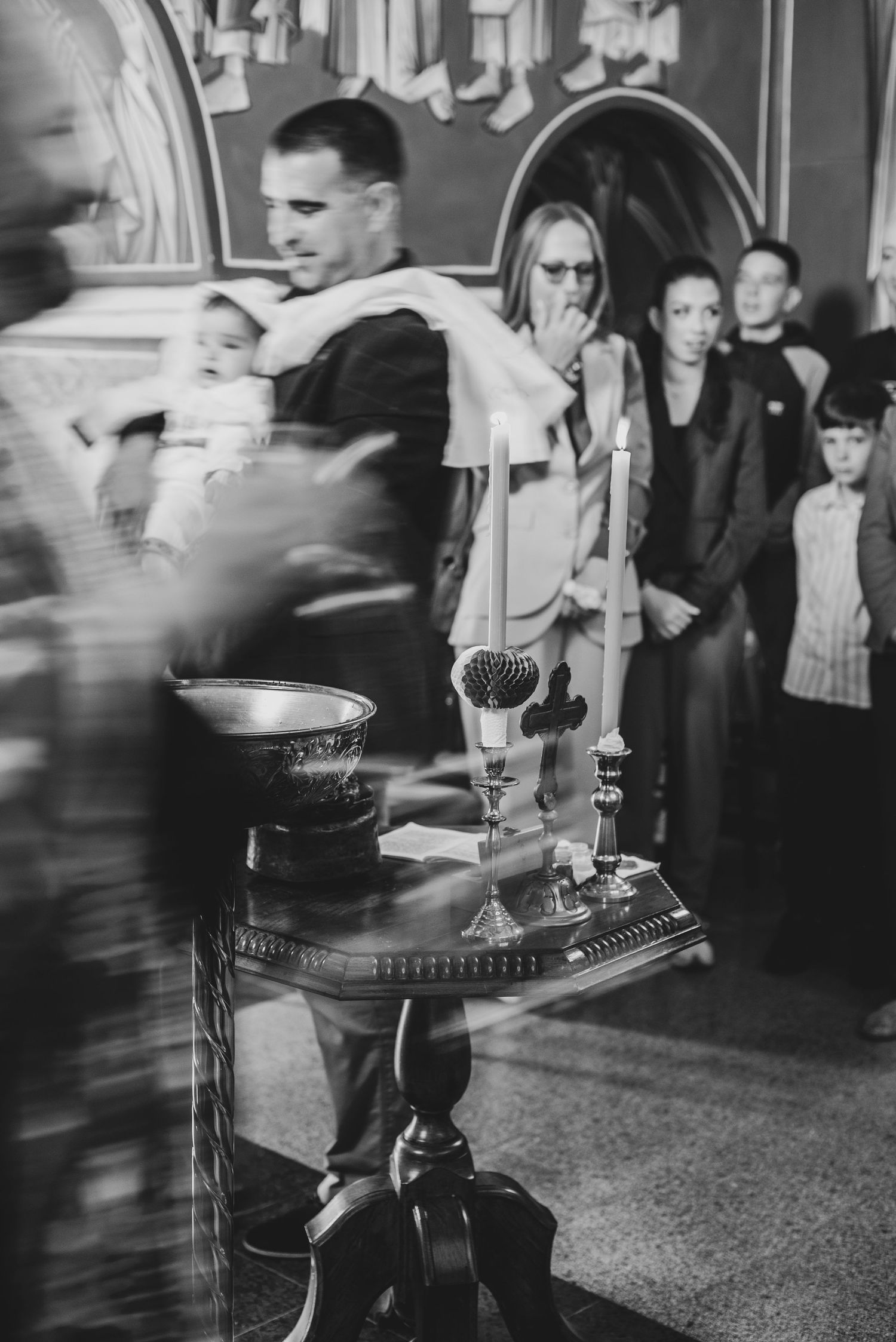 Black and white photo of a baptism ceremony with candles and a baptismal font in a church.