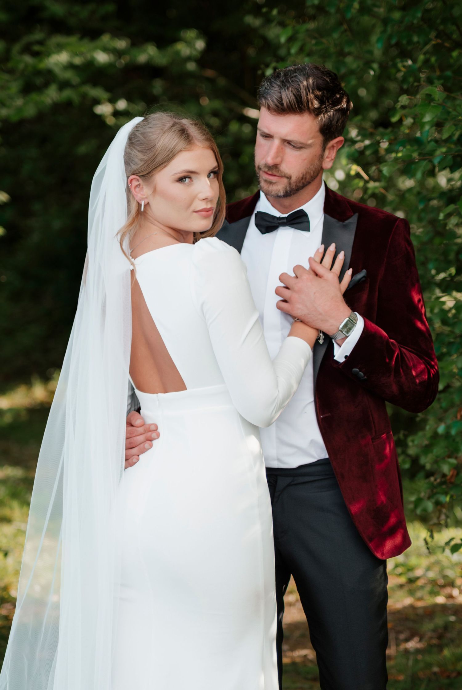 Bride in open-back gown with groom in burgundy velvet tuxedo jacket outdoors.