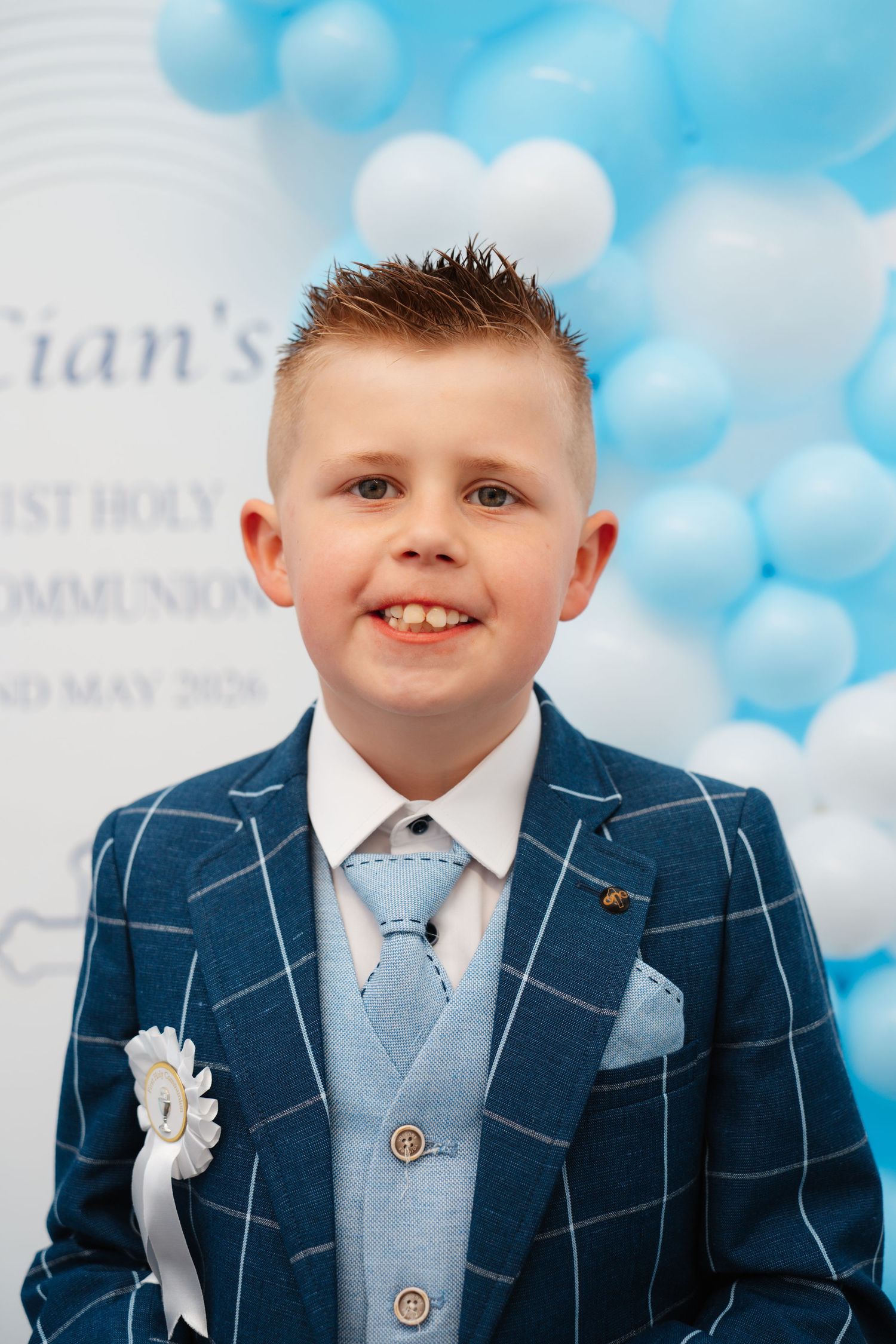Boy in blue checked suit smiling at his First Holy Communion celebration with balloon backdrop.