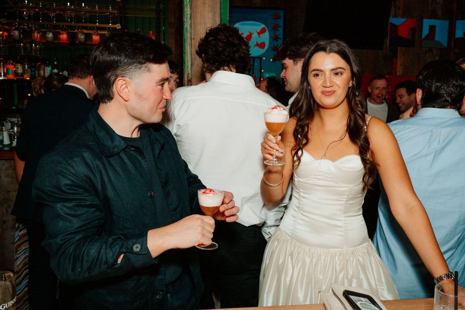 Couple in formal attire holding cocktails at a lively bar event, woman in white strapless dress smiling.