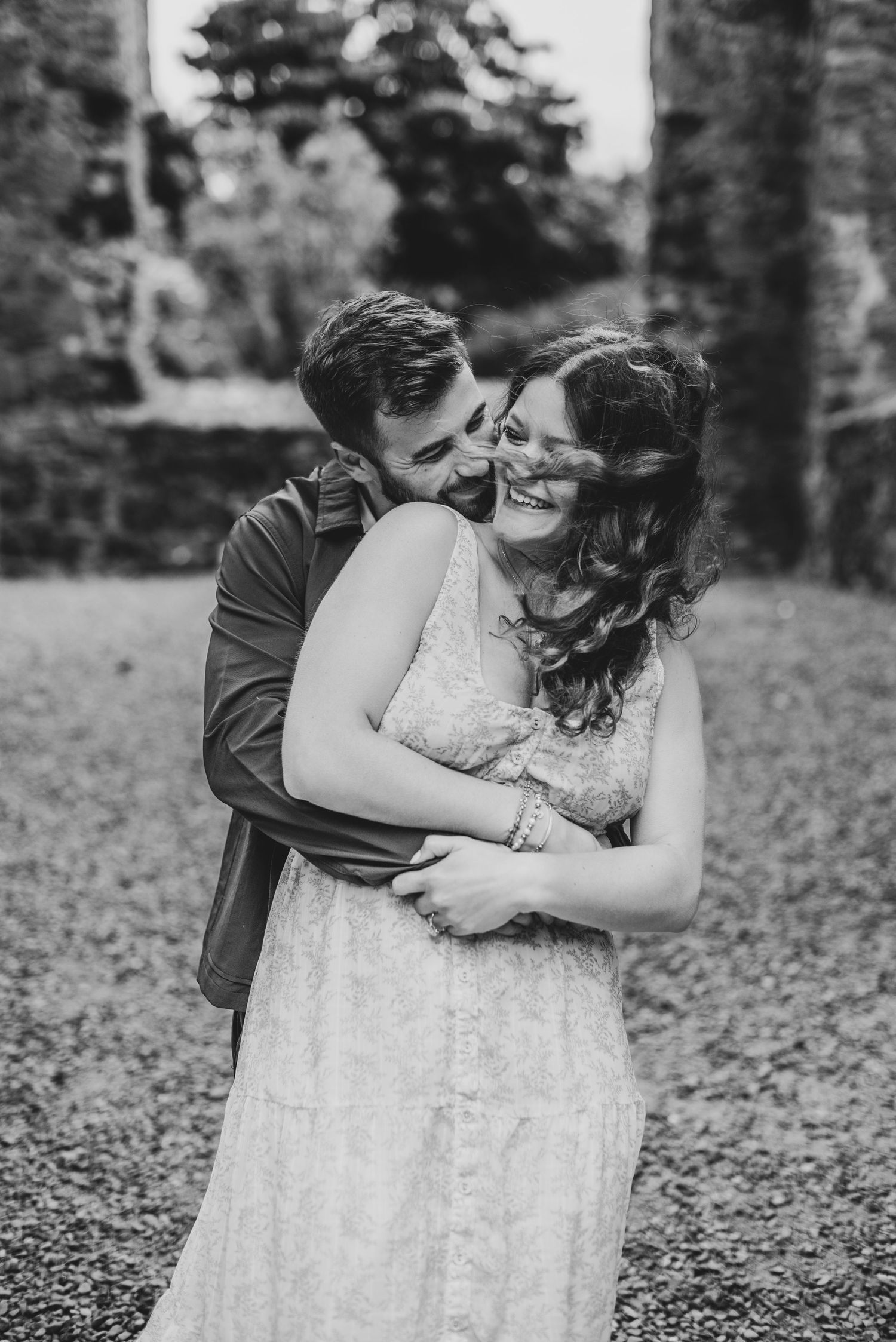 Black and white photo of a couple embracing and laughing outdoors, woman in lace dress, man in jacket.