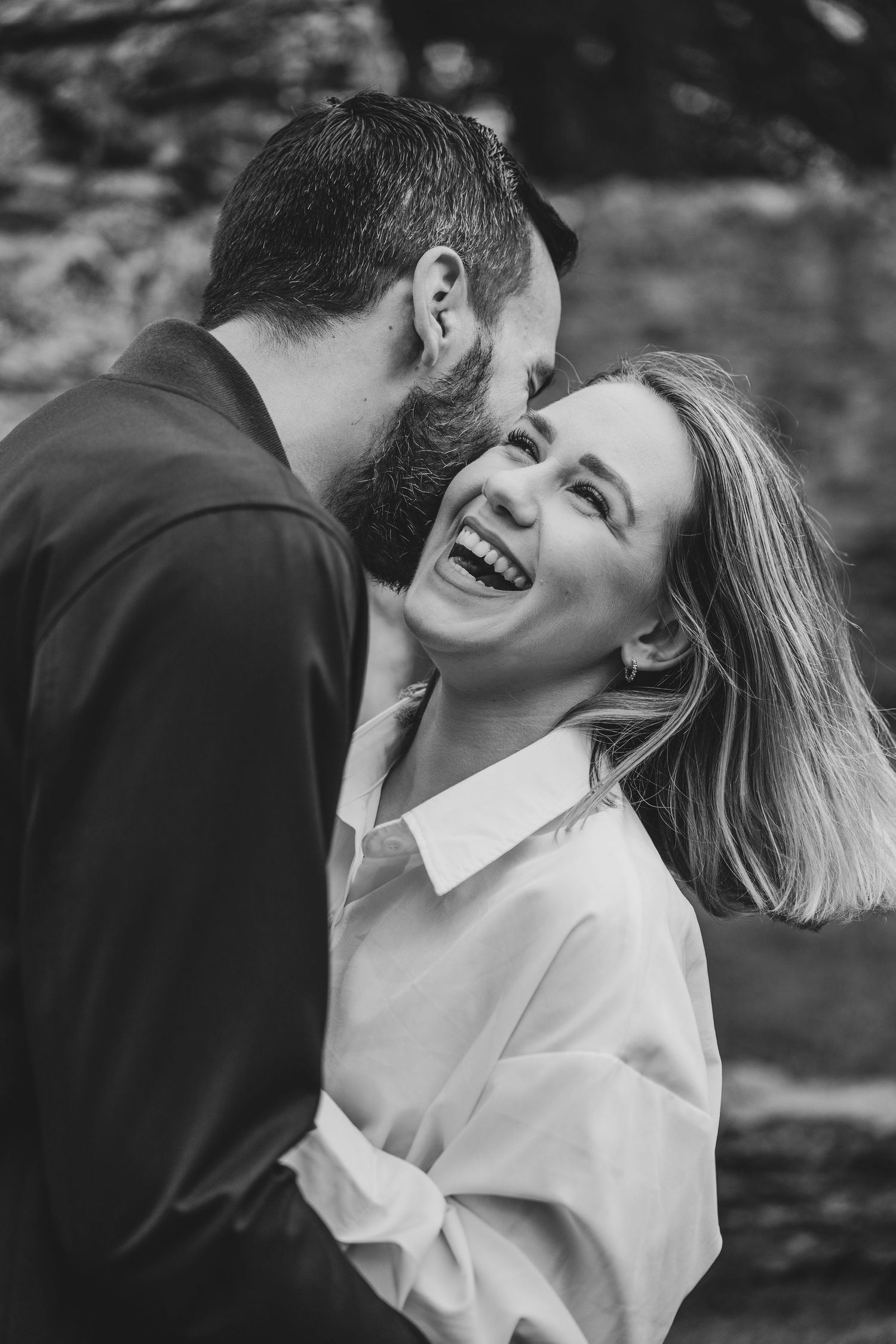 Black and white photo of a laughing woman and bearded man sharing a tender romantic moment outdoors.