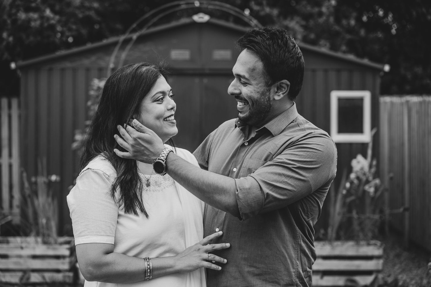 Black and white photo of a couple sharing a tender moment, man gently holding womans face while smiling at each other outdoors.