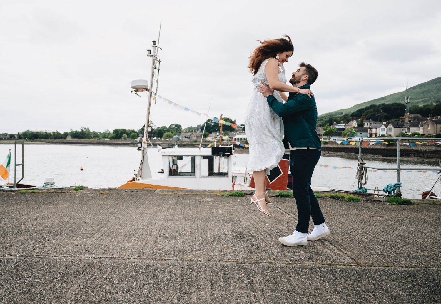 A man lifts a woman joyfully on a harbor dock, with boats and hills in the background on a cloudy day.