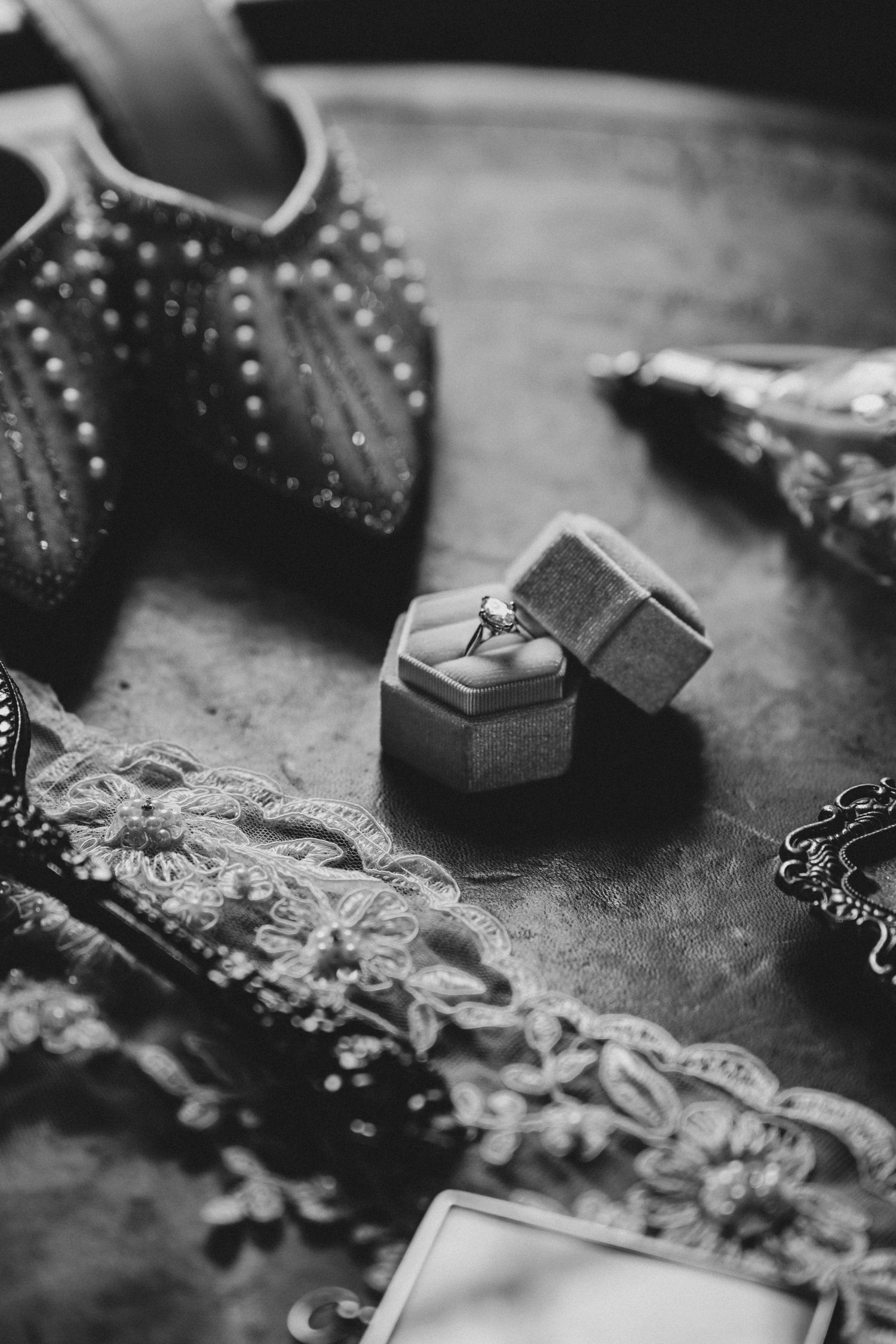Black and white photo of a diamond engagement ring in a velvet box surrounded by bridal accessories and embellished shoes.