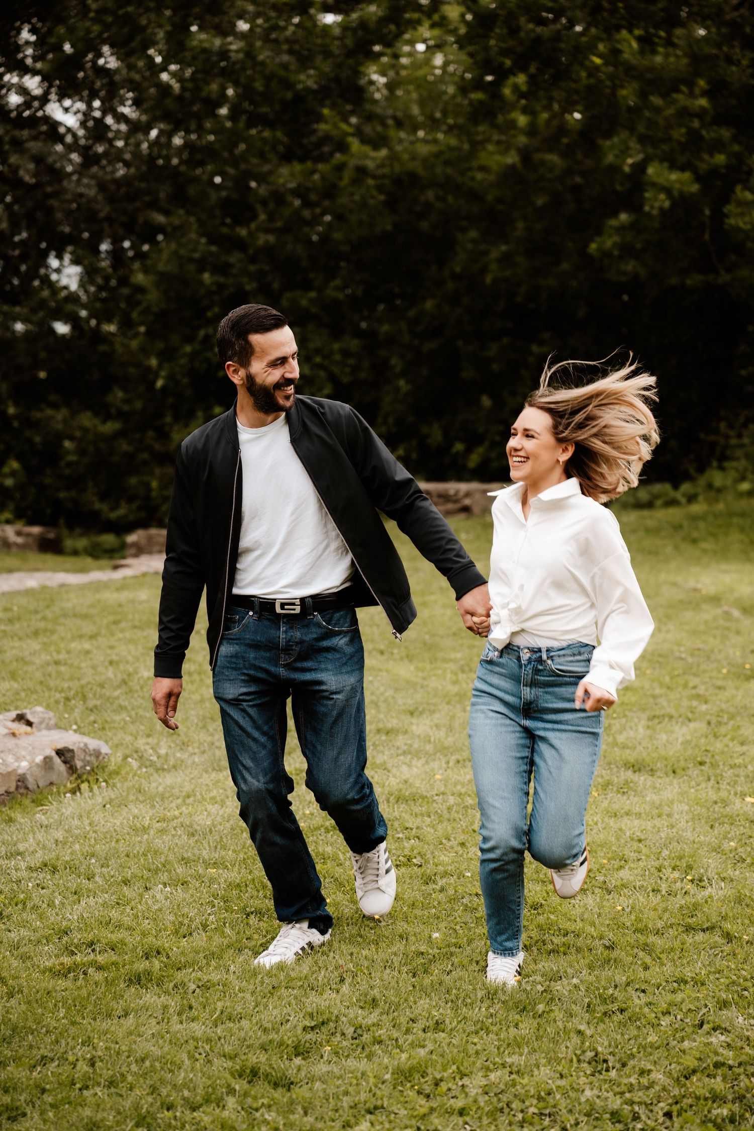 A happy couple holds hands and runs through a lush green park, smiling at each other in casual jeans and jackets.
