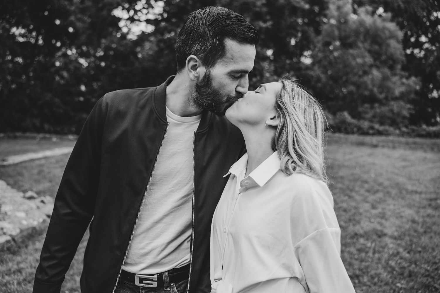 Black and white photo of a couple sharing a romantic kiss outdoors in a lush green park setting.