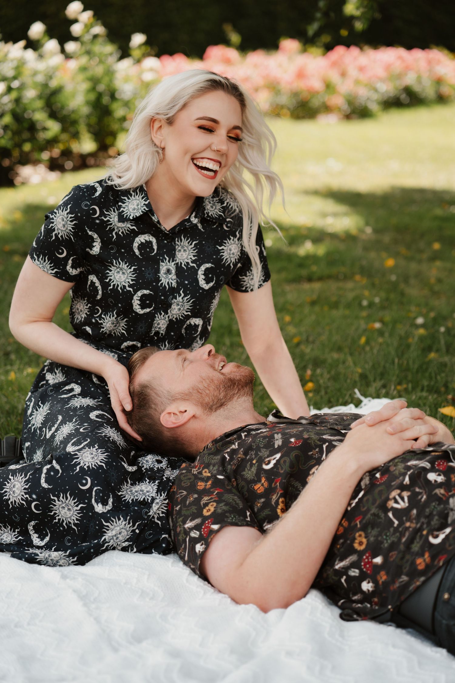 A laughing blonde woman in a celestial print dress sits beside a smiling man lying on a white blanket in a garden.