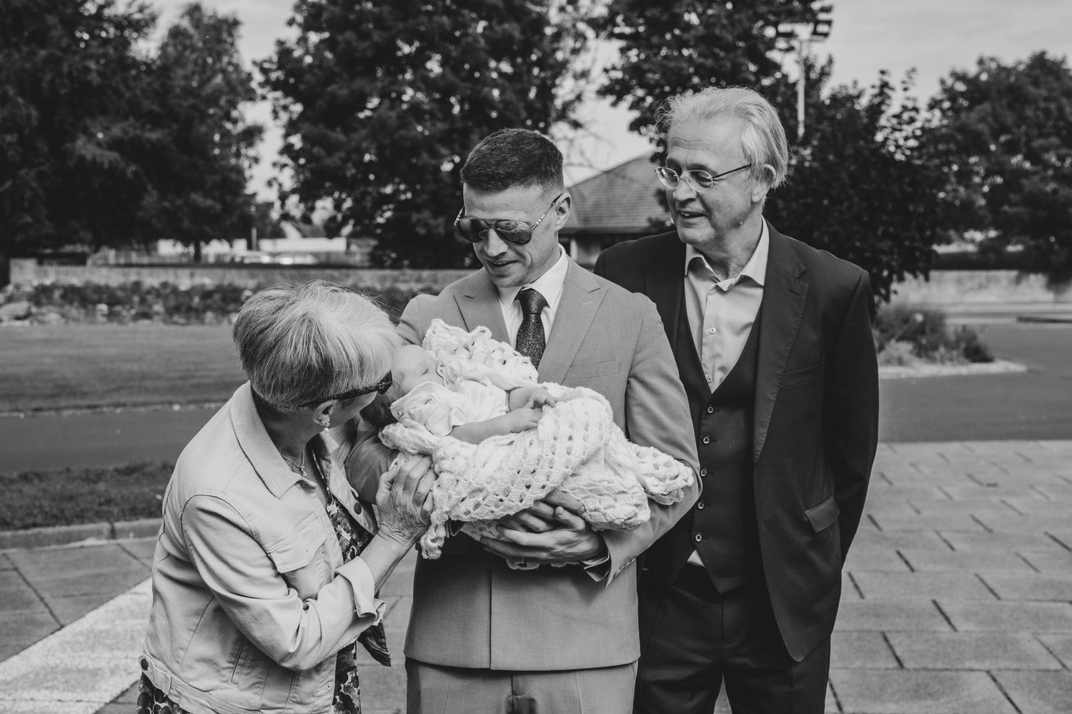 Black and white photo of a man holding a baby surrounded by smiling elderly relatives outdoors at a family gathering.