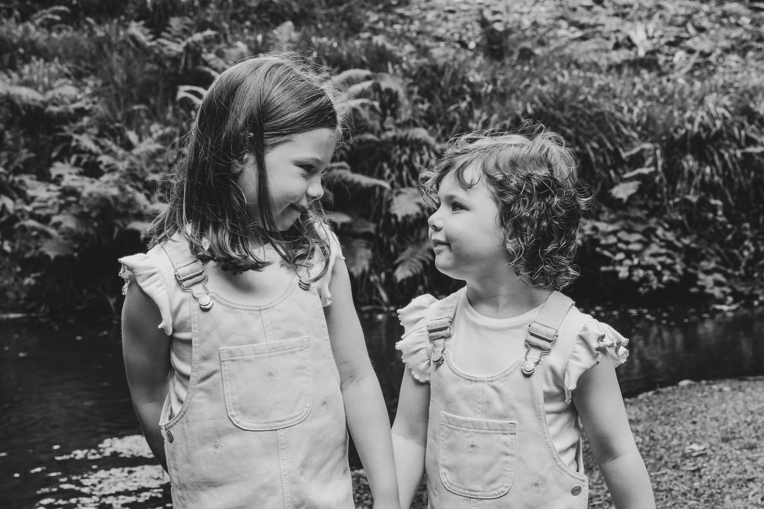 Two young girls in matching ruffle overalls hold hands and smile at each other in a black and white outdoor photo.