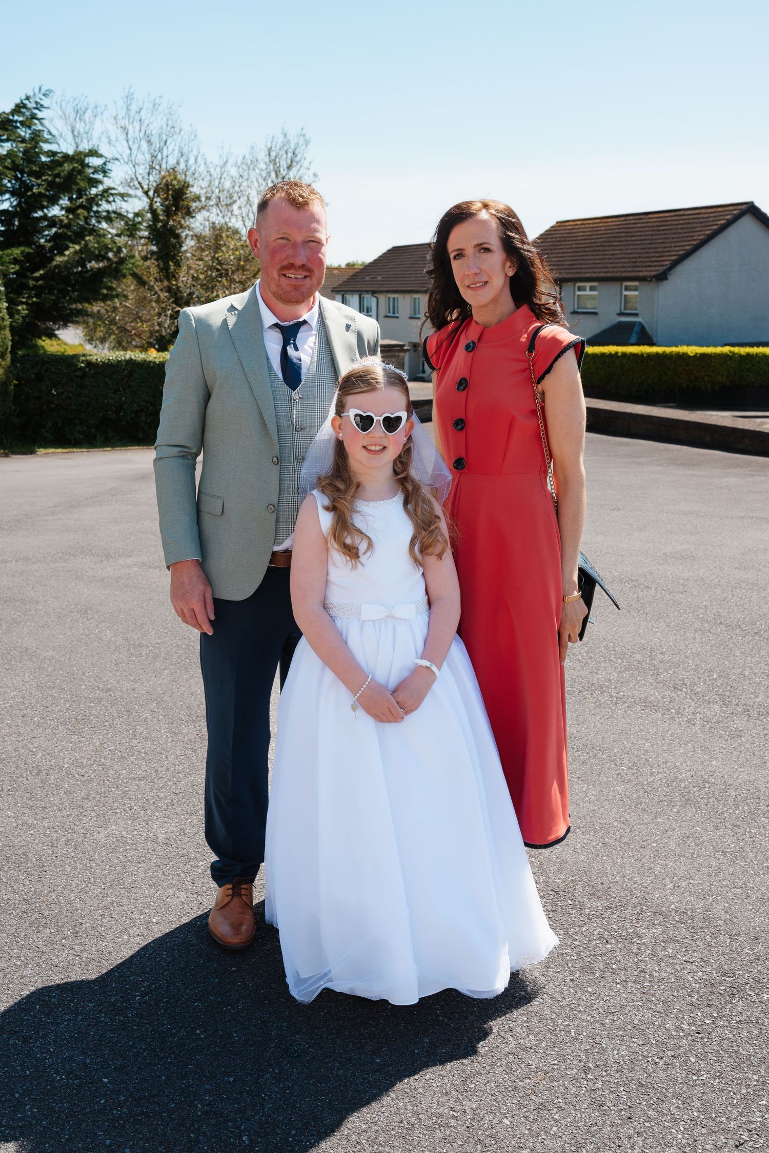 Family of three posing outdoors on sunny day, girl in white communion dress with sunglasses, parents in formal attire.