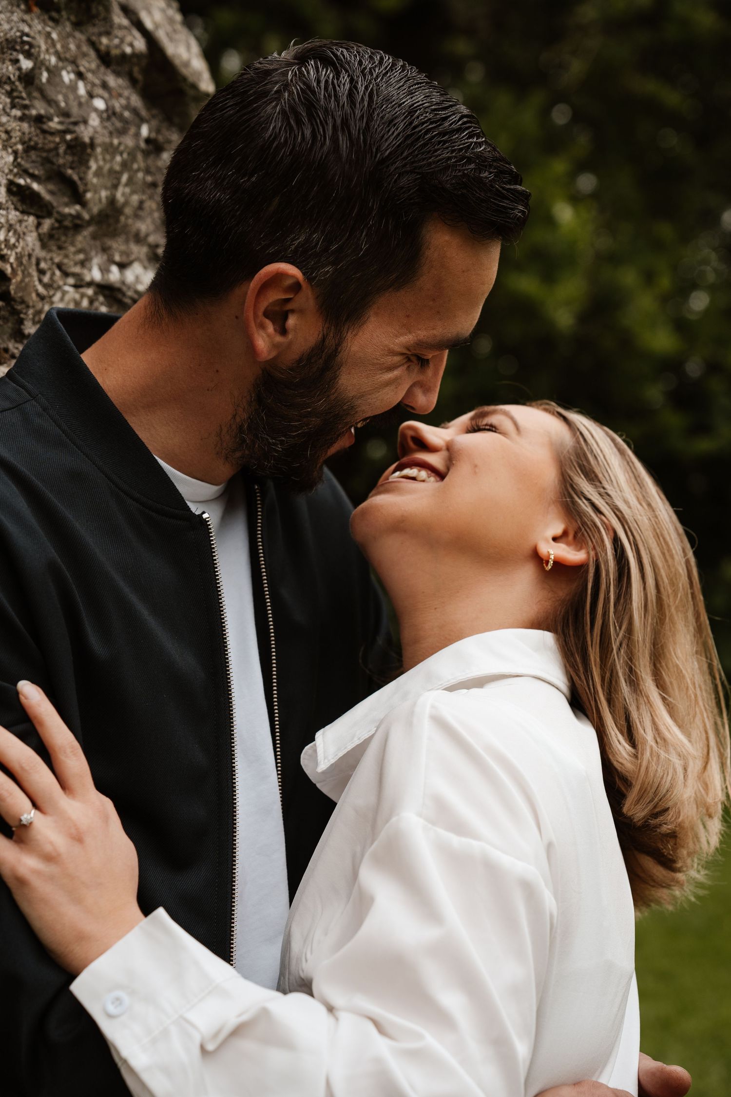 A laughing couple sharing a joyful intimate moment outdoors, the woman in white and man in black jacket nearly kissing.