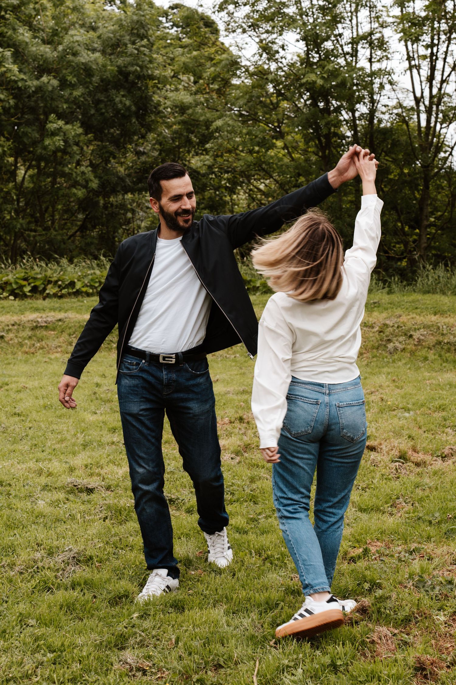 A couple dancing outdoors on green grass, man in black jacket spinning woman in white shirt and jeans.