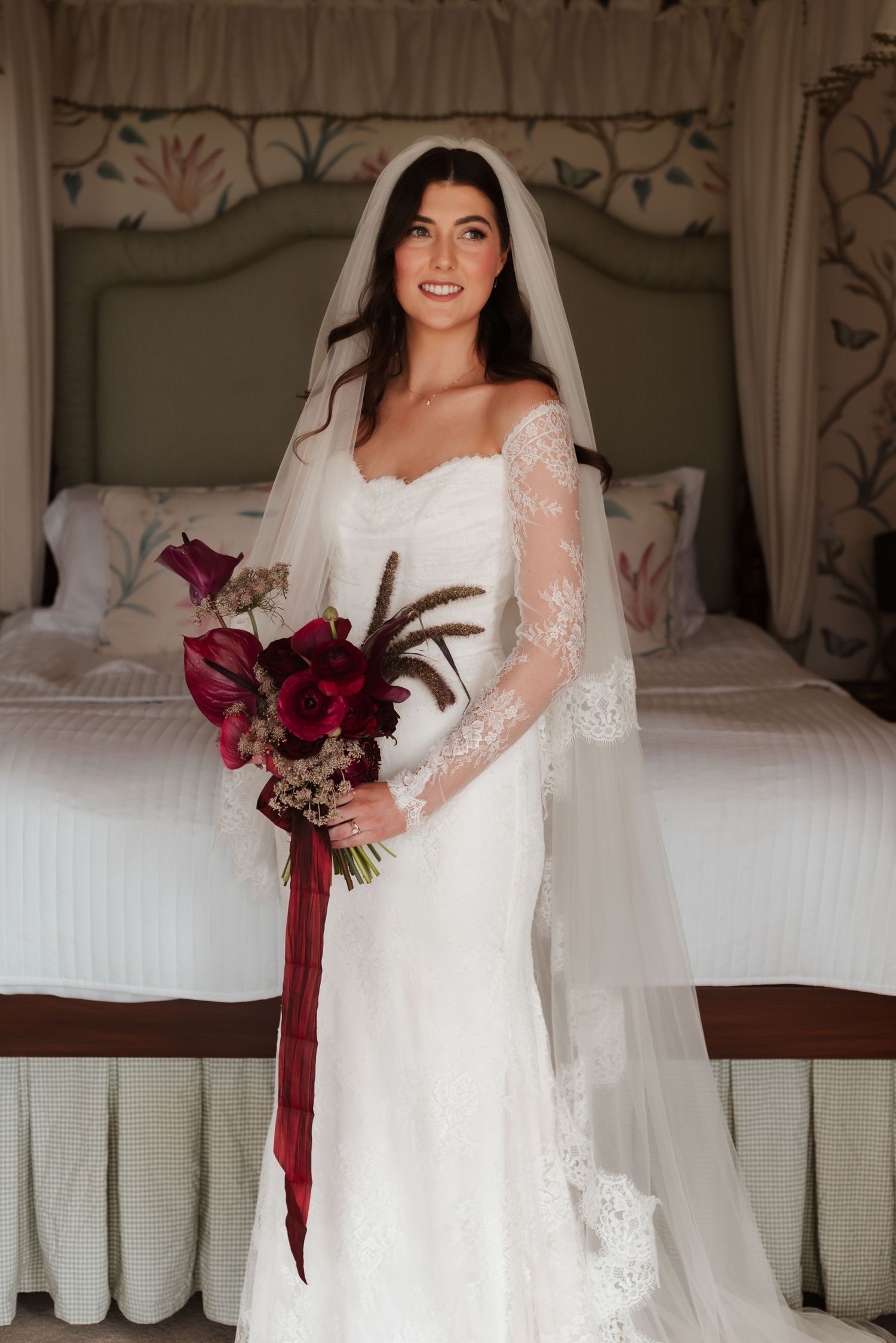 Elegant bride in lace off-shoulder gown holds burgundy bouquet with ribbon in decorated bedroom.