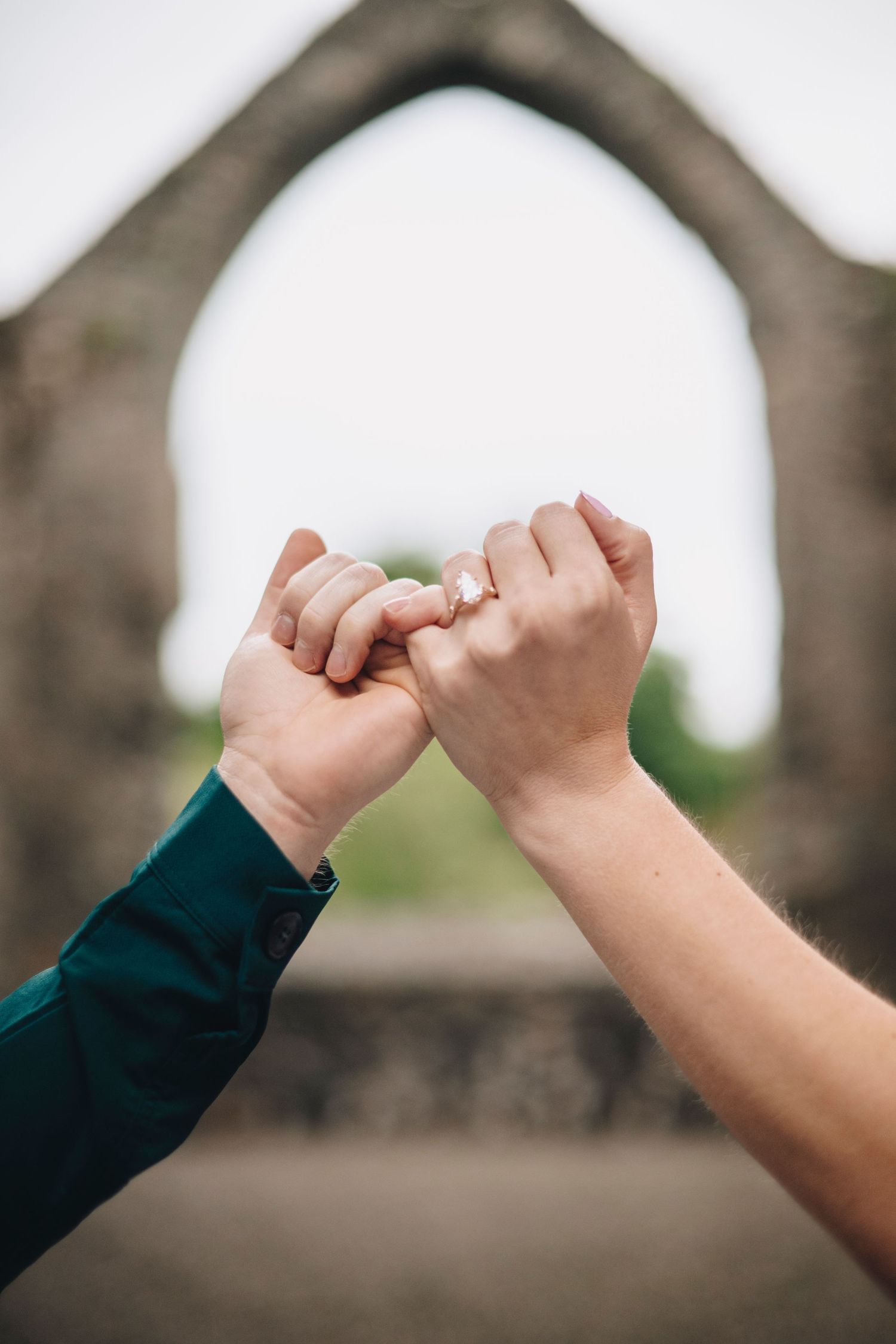 Couple holding hands with an engagement ring visible, framed by ancient stone archway ruins in the background.