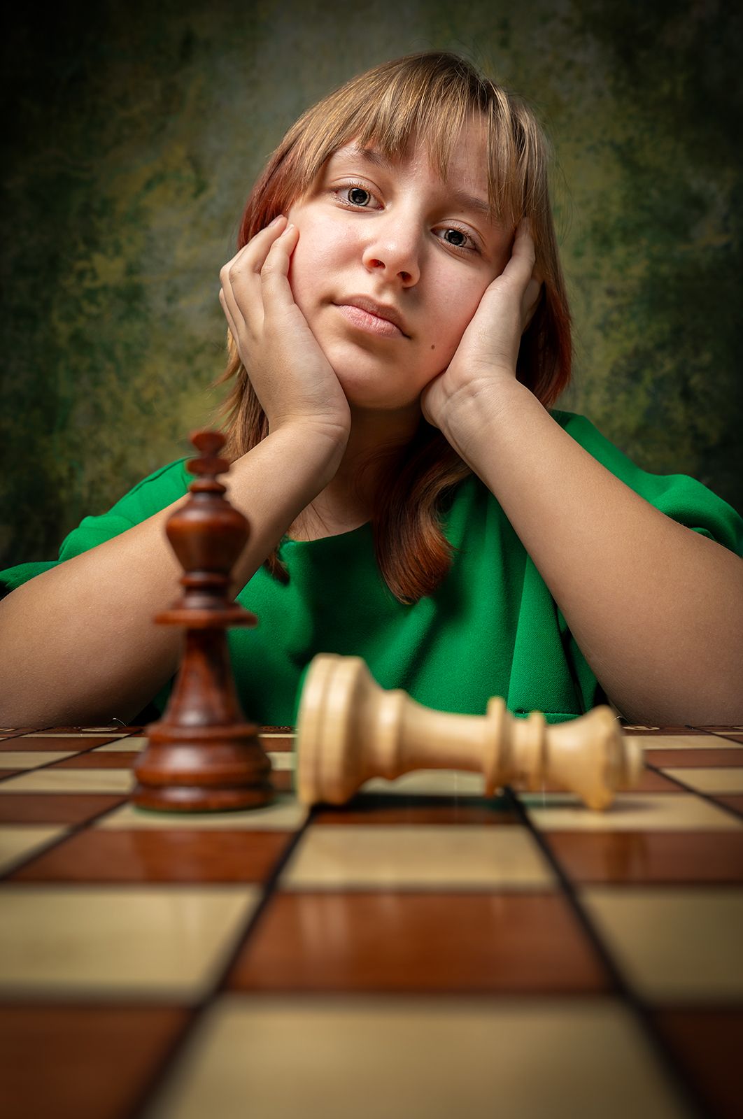 Young girl in green shirt rests chin on hands, contemplating a chess board with fallen white king piece.