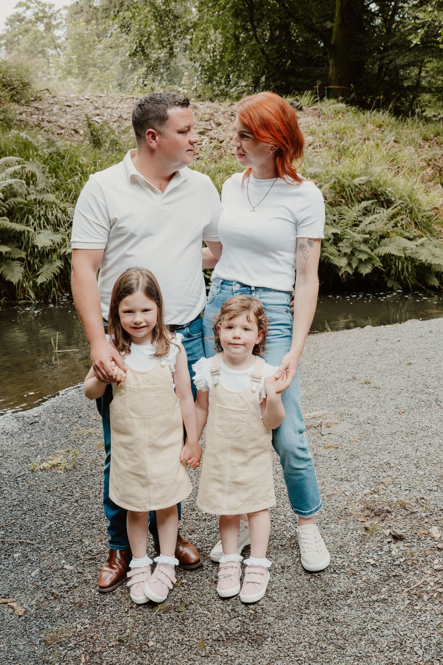 Family of four posing outdoors near a stream, parents in white tops and jeans, two young girls in matching beige pinafore dresses.