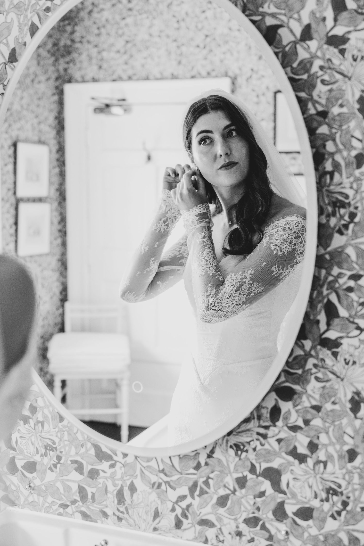 Bride in lace wedding dress adjusting earring reflected in ornate oval mirror, black and white photo.