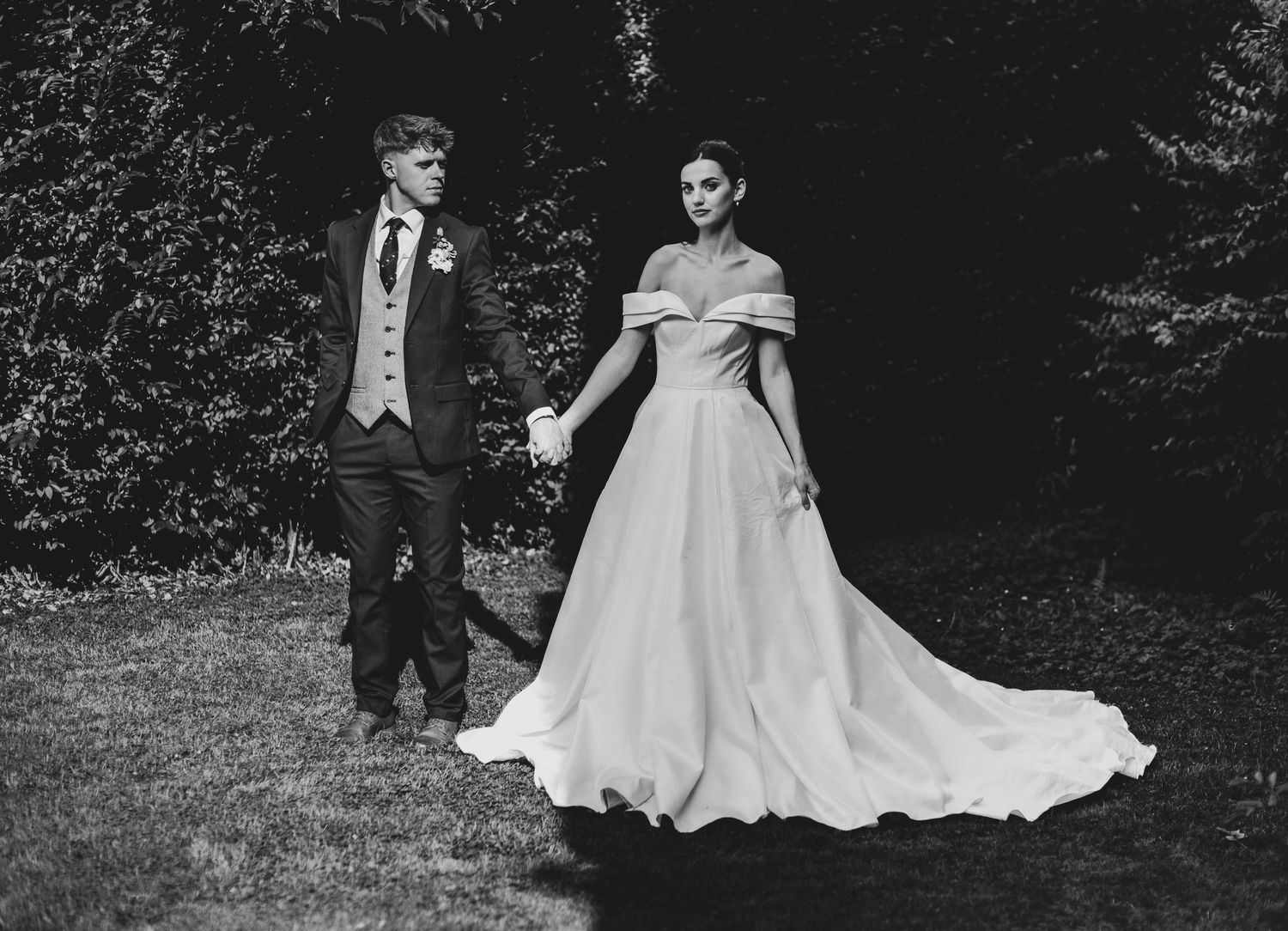 Black and white wedding portrait of couple holding hands in lush garden, bride in off-shoulder gown.