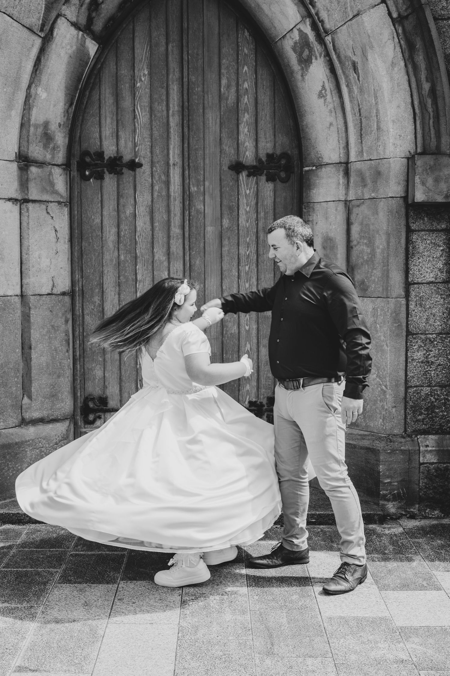 Black and white photo of a man spinning a girl in a white dress outside a stone building with a wooden arched door.