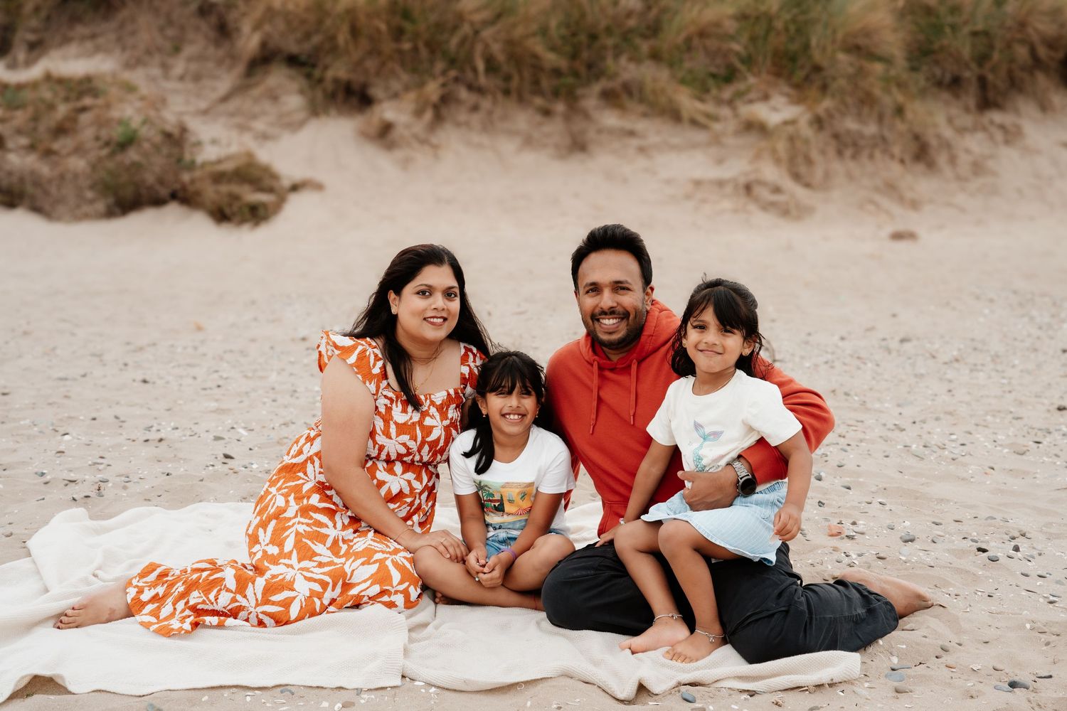 Happy family of four sitting on a beach blanket on sandy shores, parents and two young children smiling together.