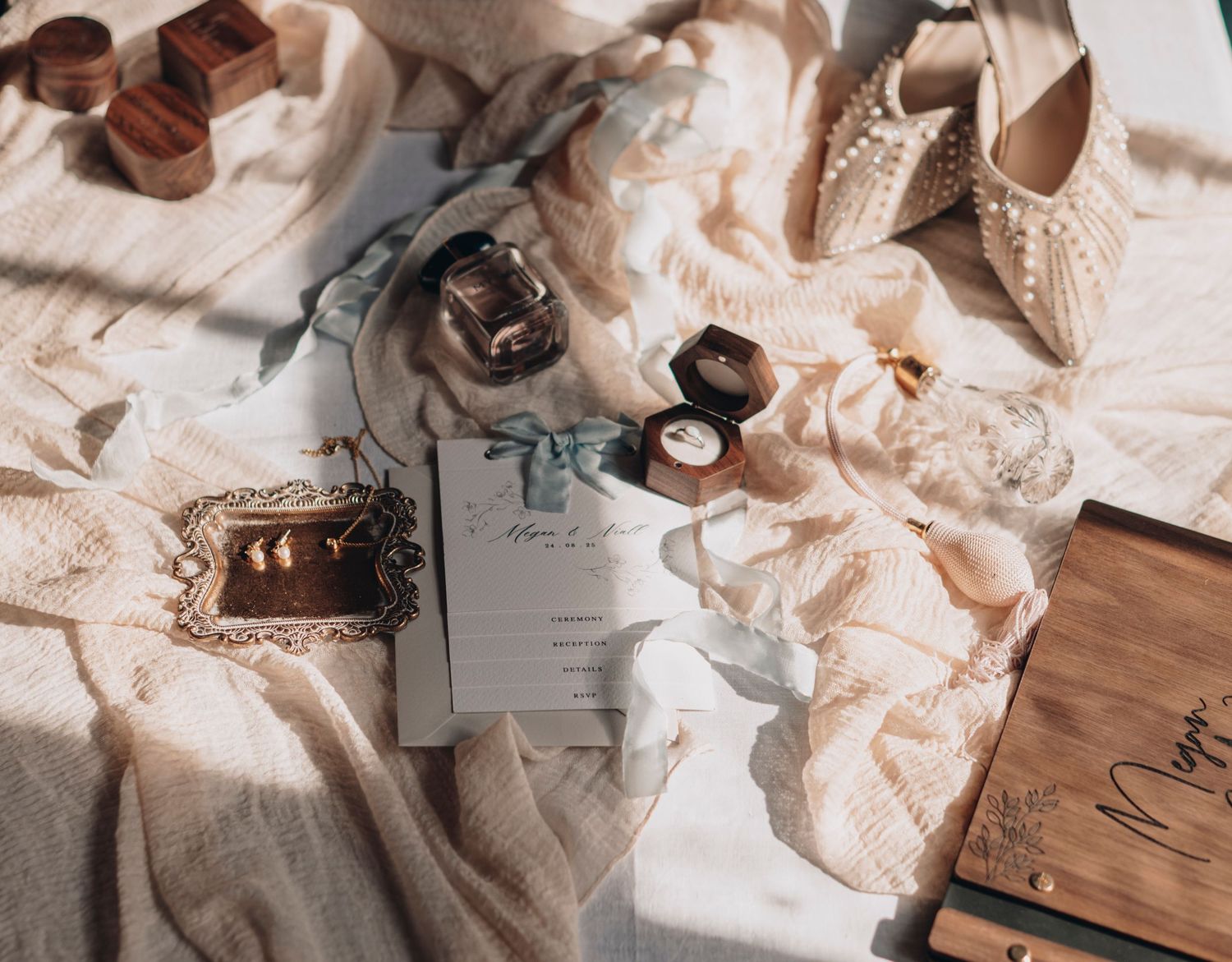 Elegant wedding flat lay with bridal shoes, ring boxes, invitation, and accessories on soft cream fabric.