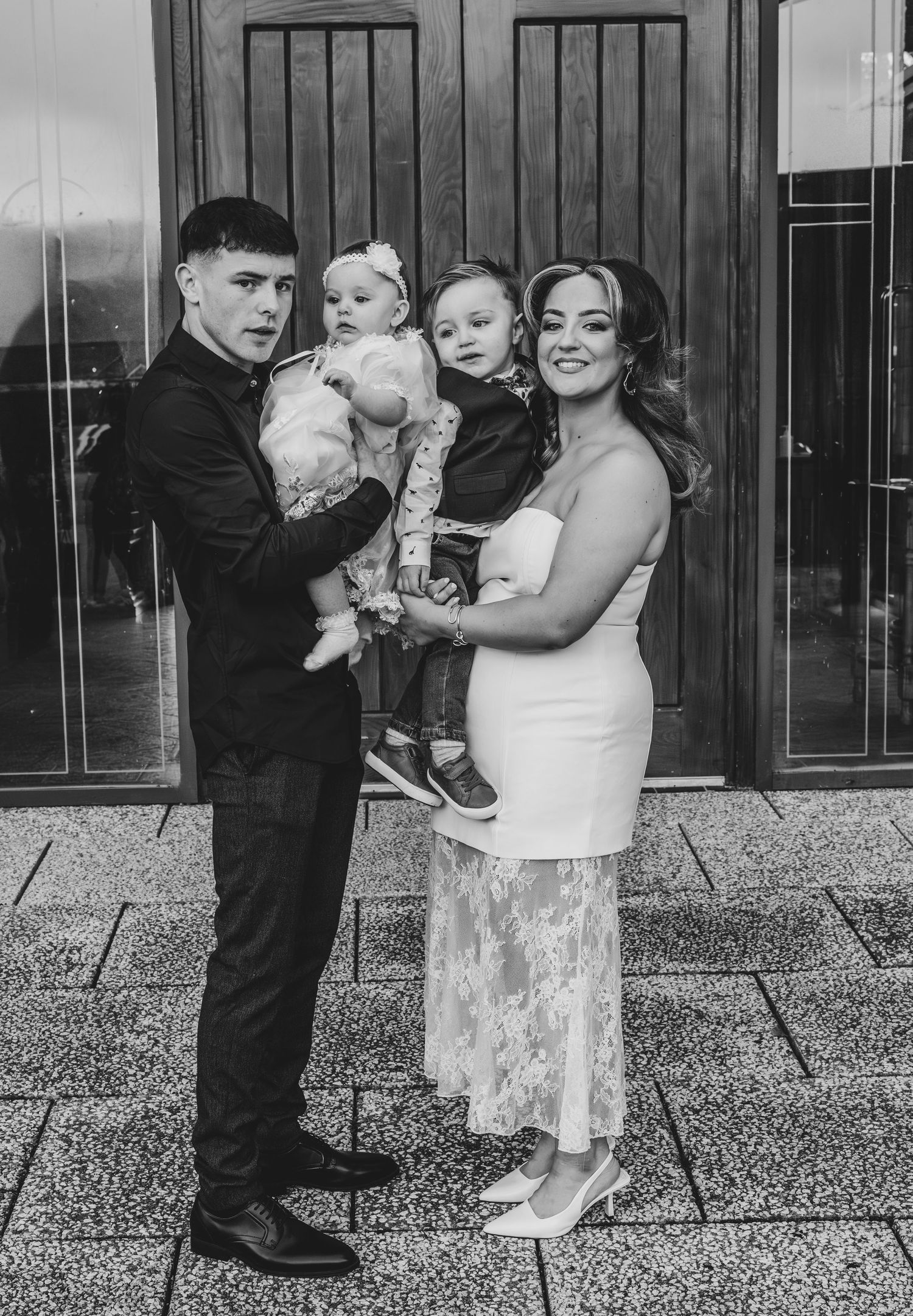 Black and white family portrait of parents holding two young children outside venue.