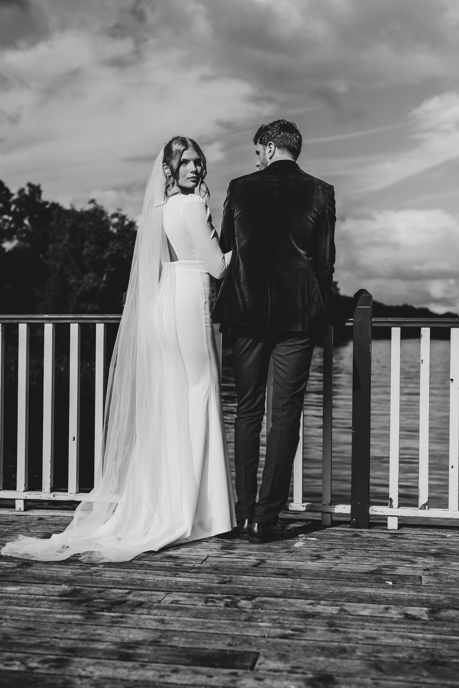 Black and white photo of bride and groom standing at railing on a lakeside deck.