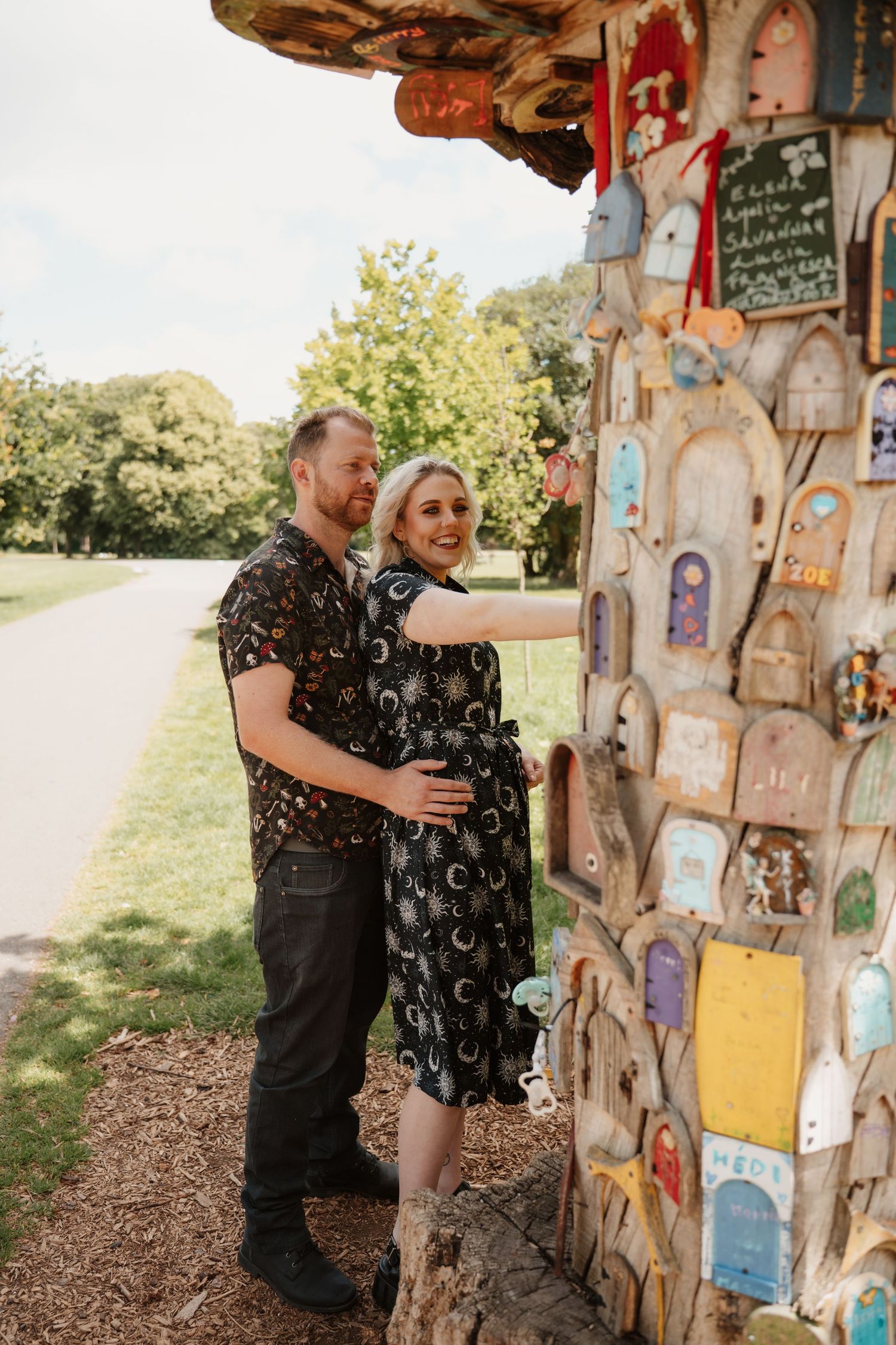 Smiling couple embracing beside a colorful wooden birdhouse tree display in a sunny park setting.