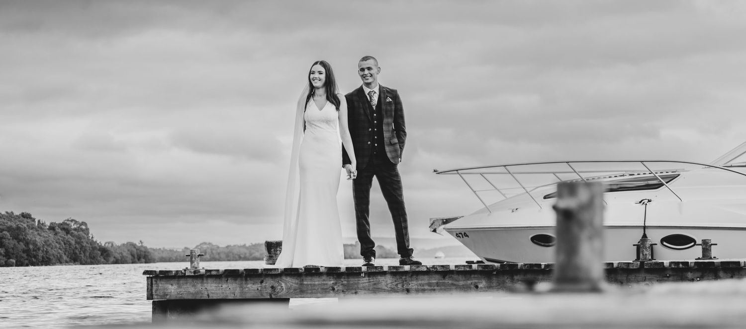Black and white photo of a bride and groom standing on a dock by a yacht on a cloudy wedding day.