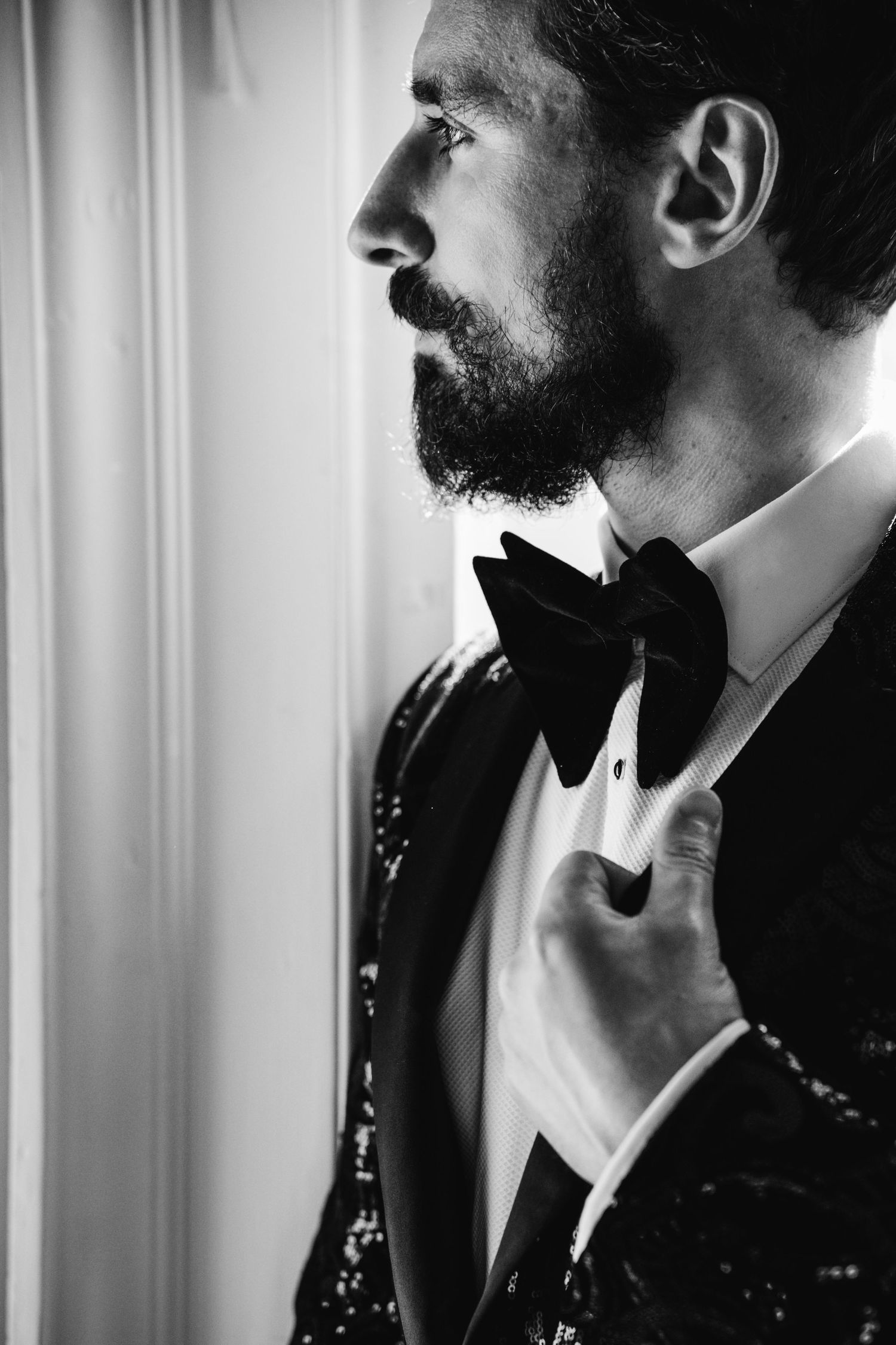 Dapper groom in sequin tuxedo jacket adjusting black velvet bow tie by window in black and white portrait.