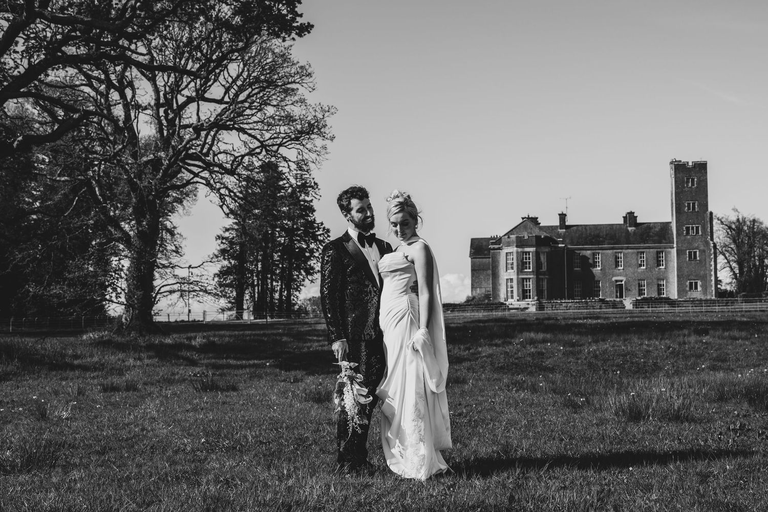 Black and white photo of a bride and groom posing in front of a grand estate on their wedding day.