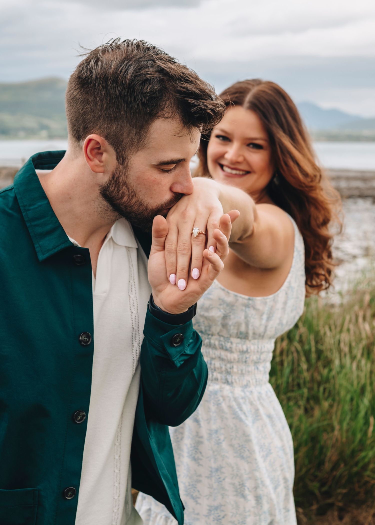 Man kissing woman's hand showing engagement ring outdoors with scenic lake and mountain backdrop.