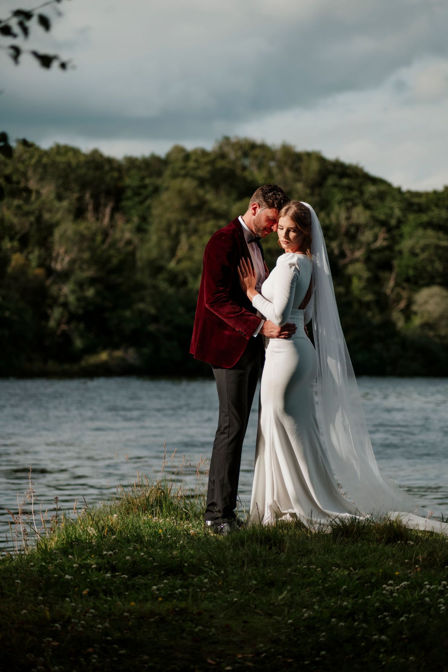 Bride in white gown with long veil embracing groom in burgundy velvet jacket by a scenic lake.