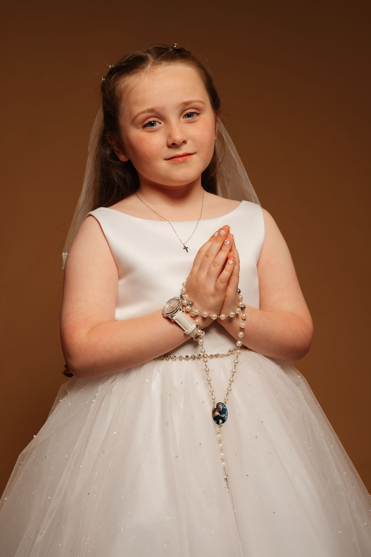 Young girl in white First Communion dress and veil, holding rosary beads in prayer position.
