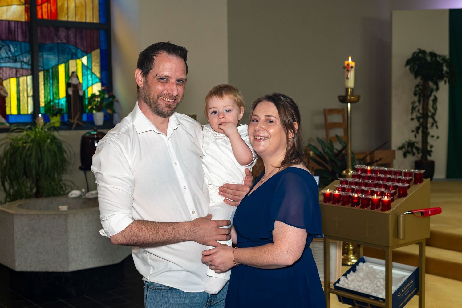 Family posing together in a church, man holding toddler beside woman in navy dress, stained glass window background.