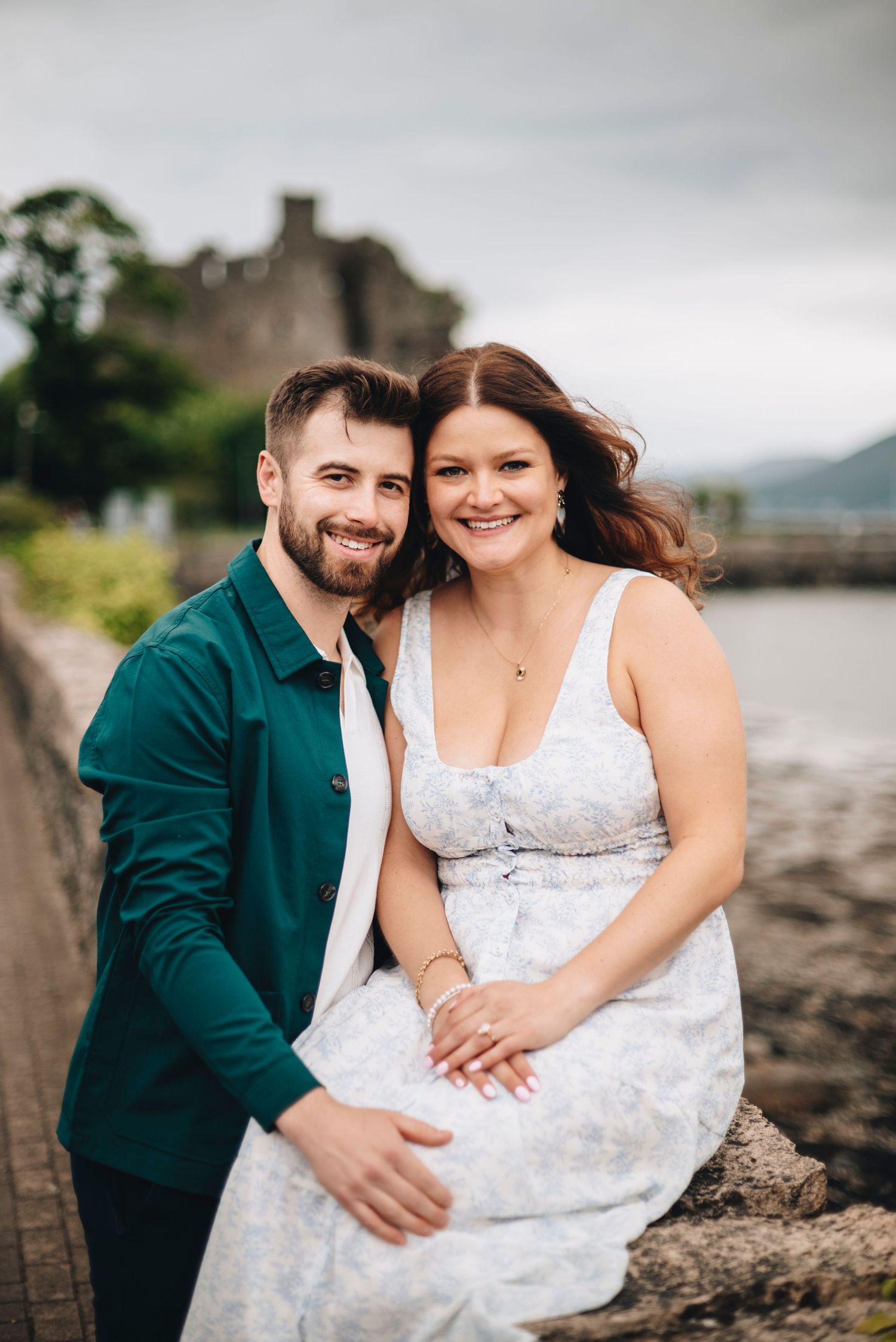 Smiling couple posing together by a seaside wall with a castle ruin in the background on a cloudy day.