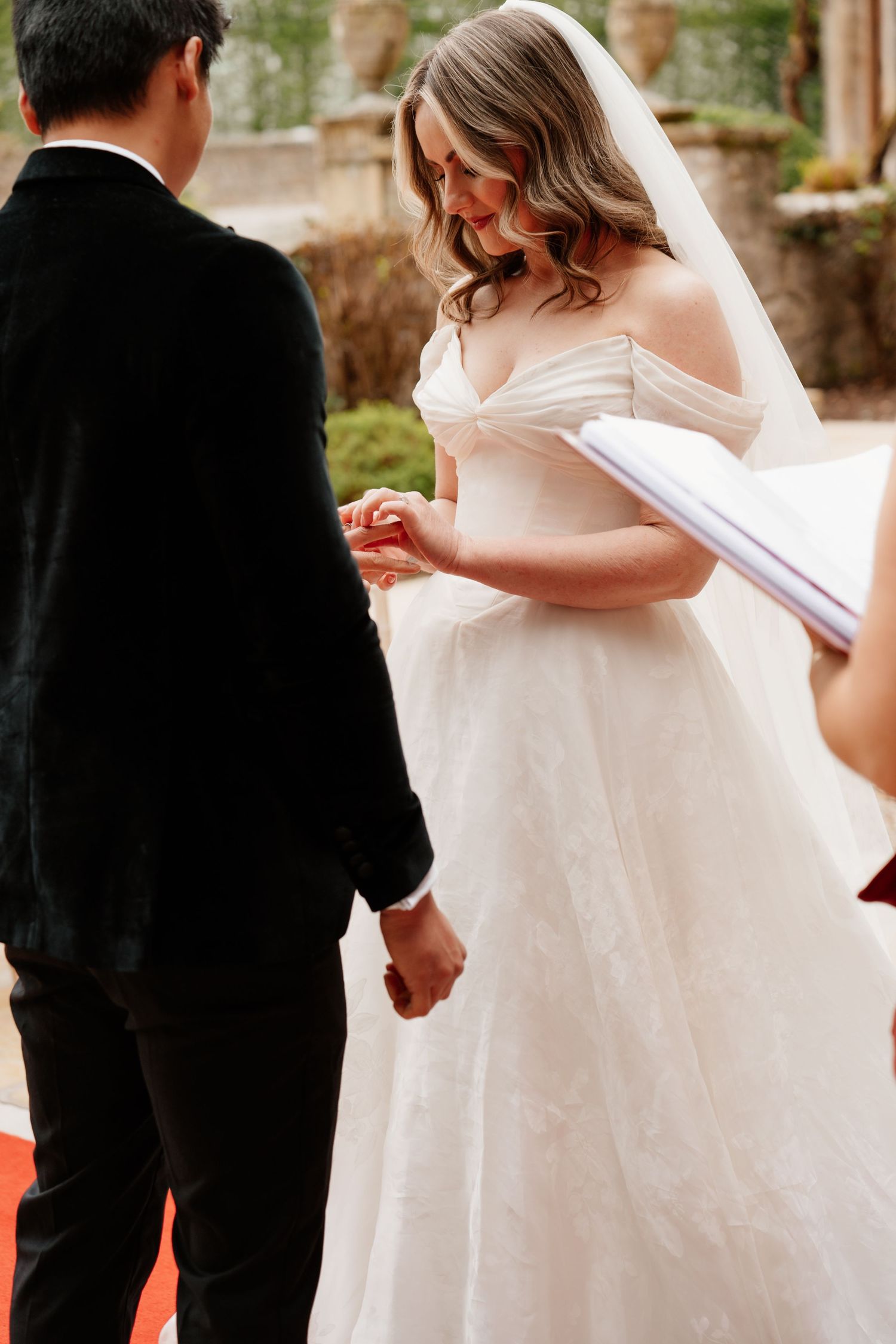 Bride in off-shoulder lace ballgown places ring on groom's finger during outdoor wedding ceremony.