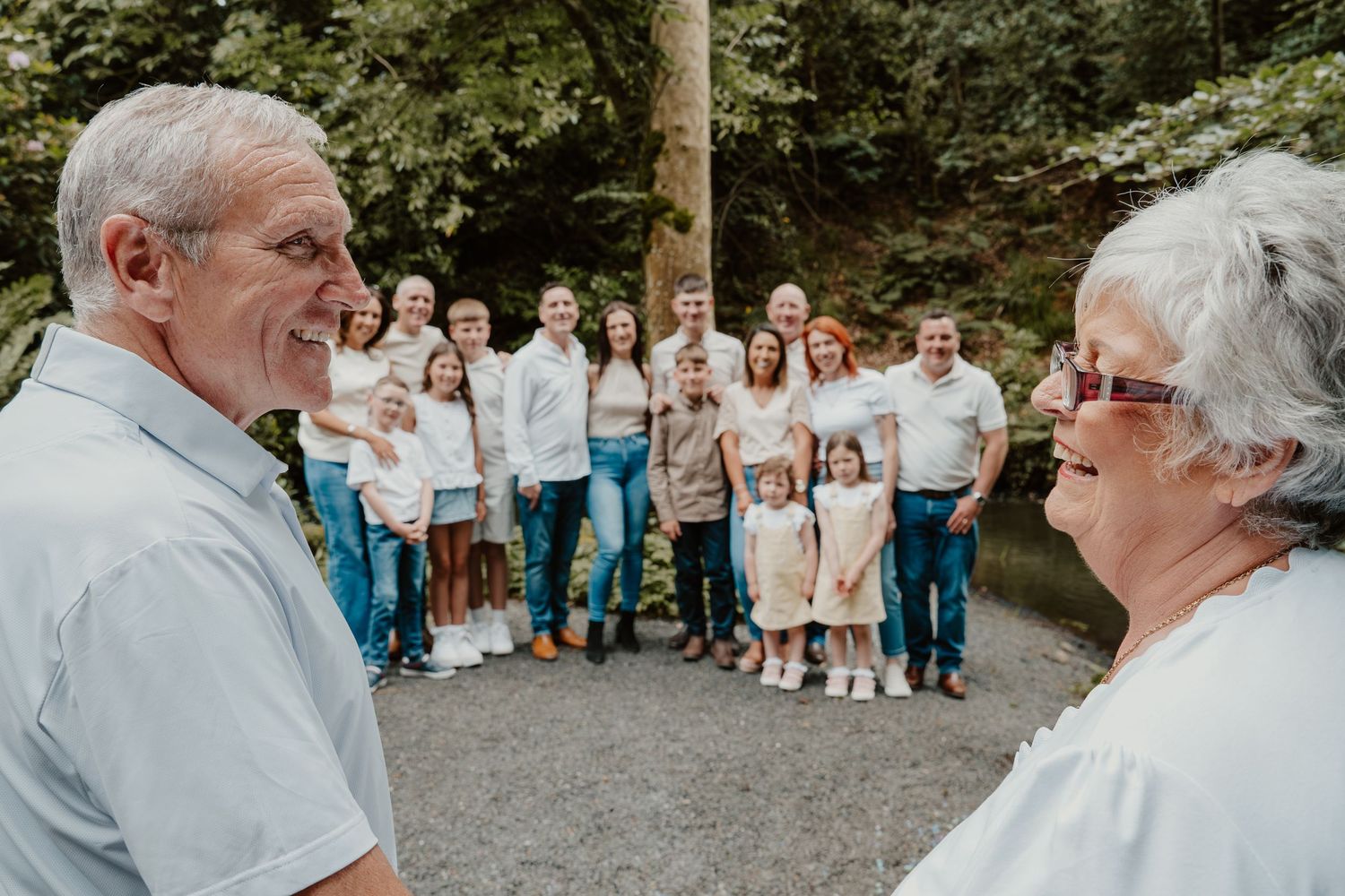 Elderly couple smiling at each other with a large multigenerational family group posed outdoors in a lush green forest setting.
