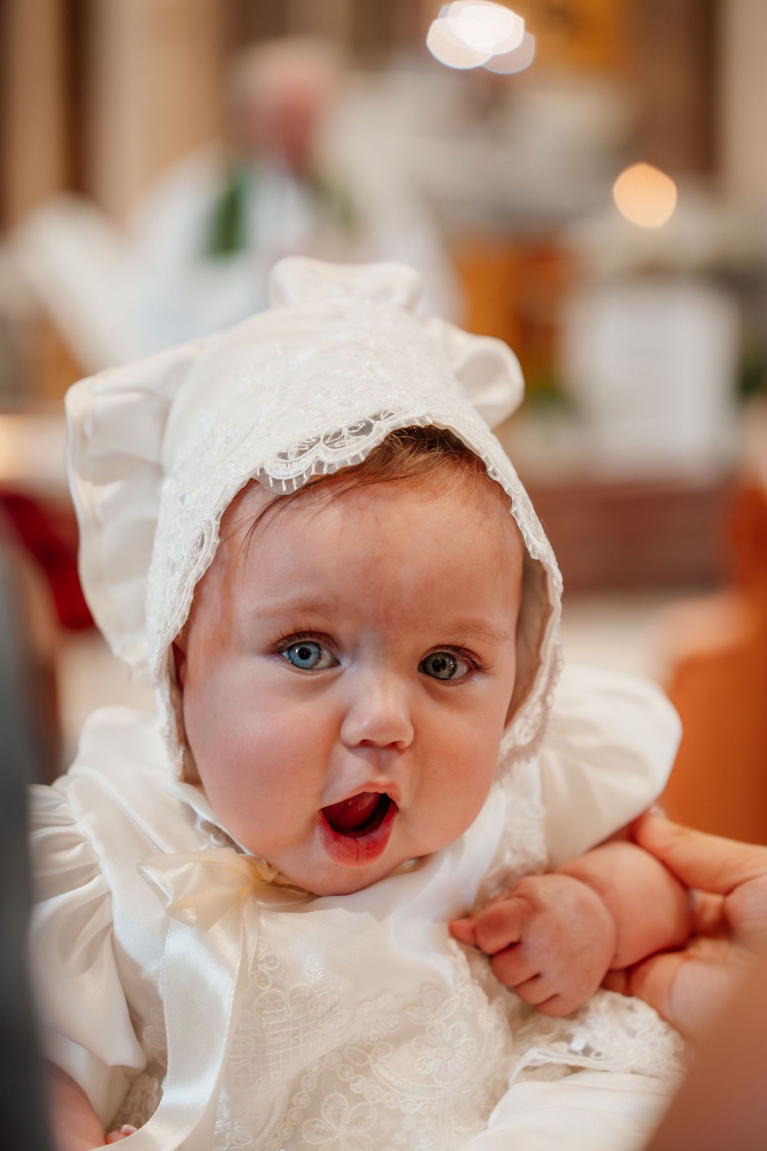 Adorable baby in white lace christening gown and bonnet with blue eyes and open mouth at baptism ceremony.