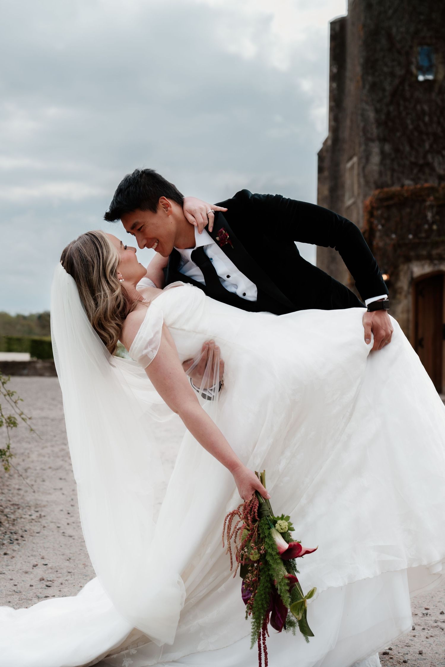 Bride in white gown holds bouquet as groom lifts her in a romantic outdoor wedding photo near a stone building.