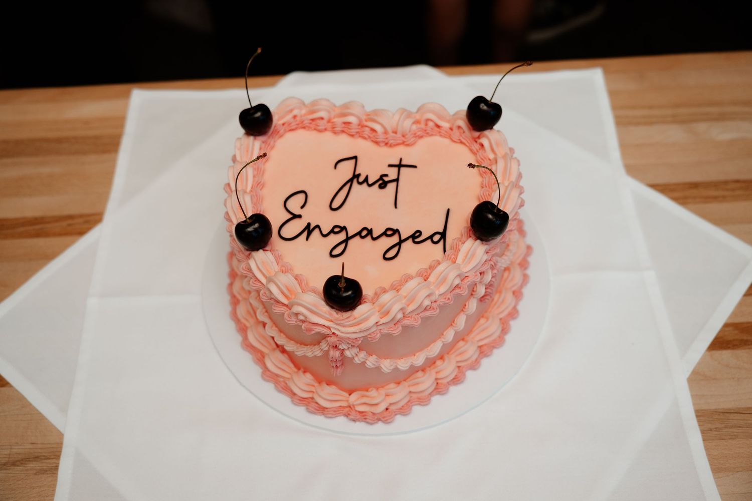 Heart-shaped pink engagement cake with cherries, decorated with Just Engaged script frosting on white plate.