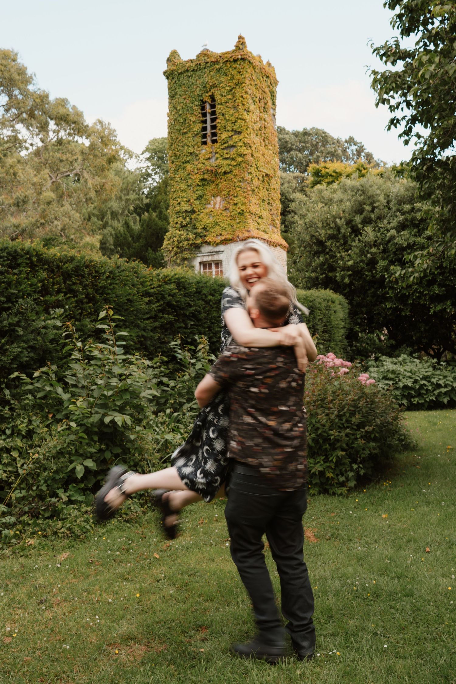 Joyful couple embracing in a green garden with an ivy-covered tower in the background.